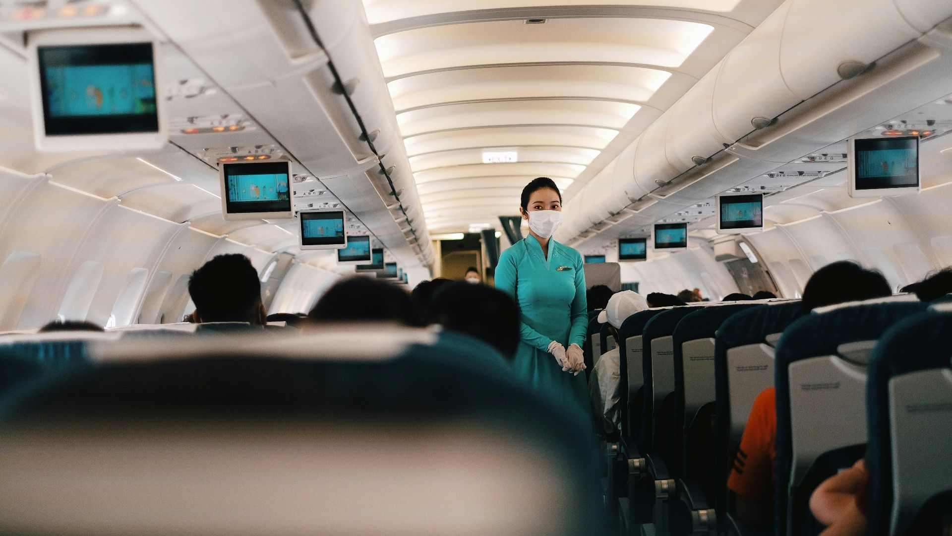 Flight attendant wearing a face mask attending to passengers in an airplane cabin.