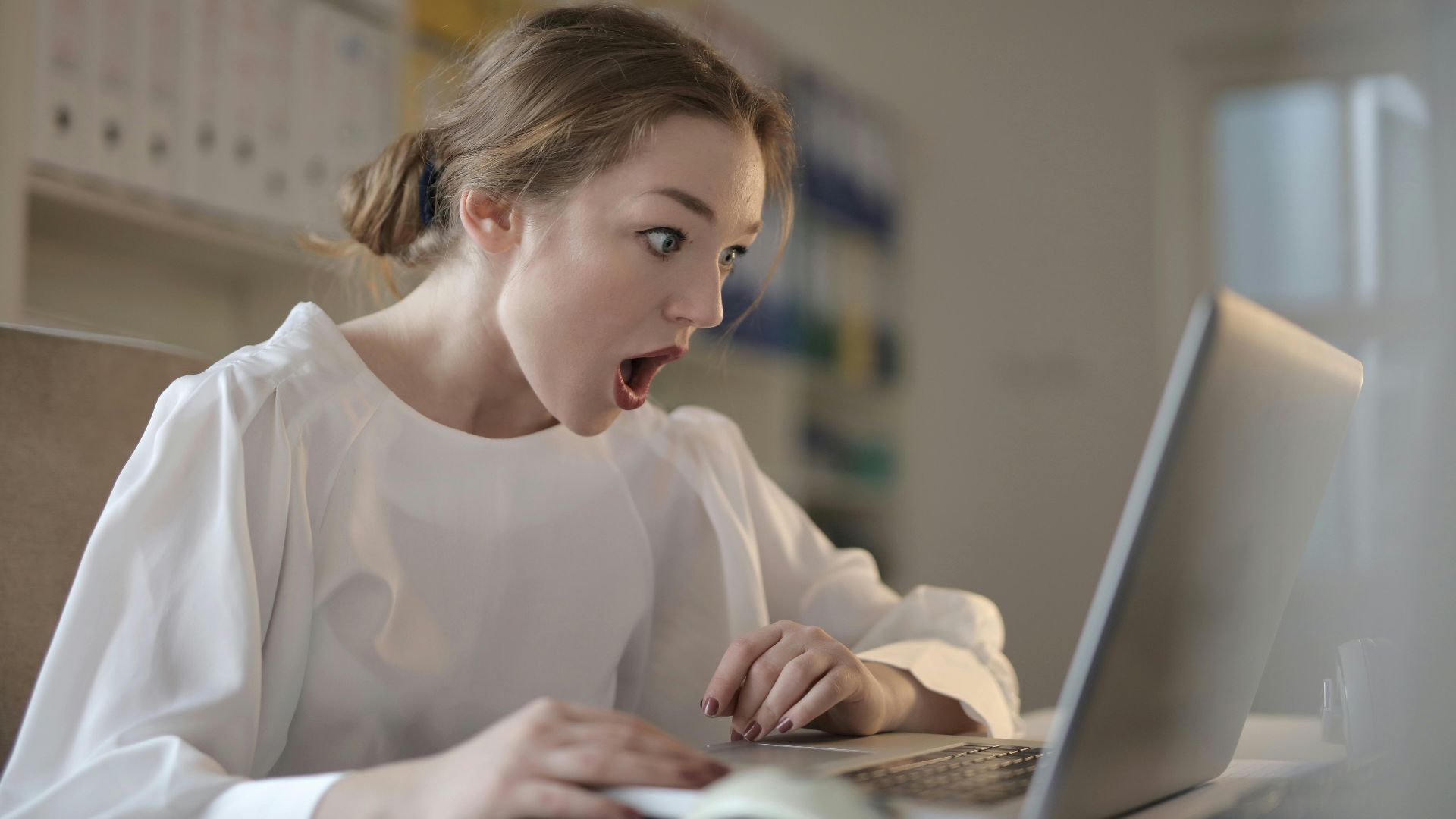 Surprised woman sitting at desk with laptop indoors, expressing amazement.