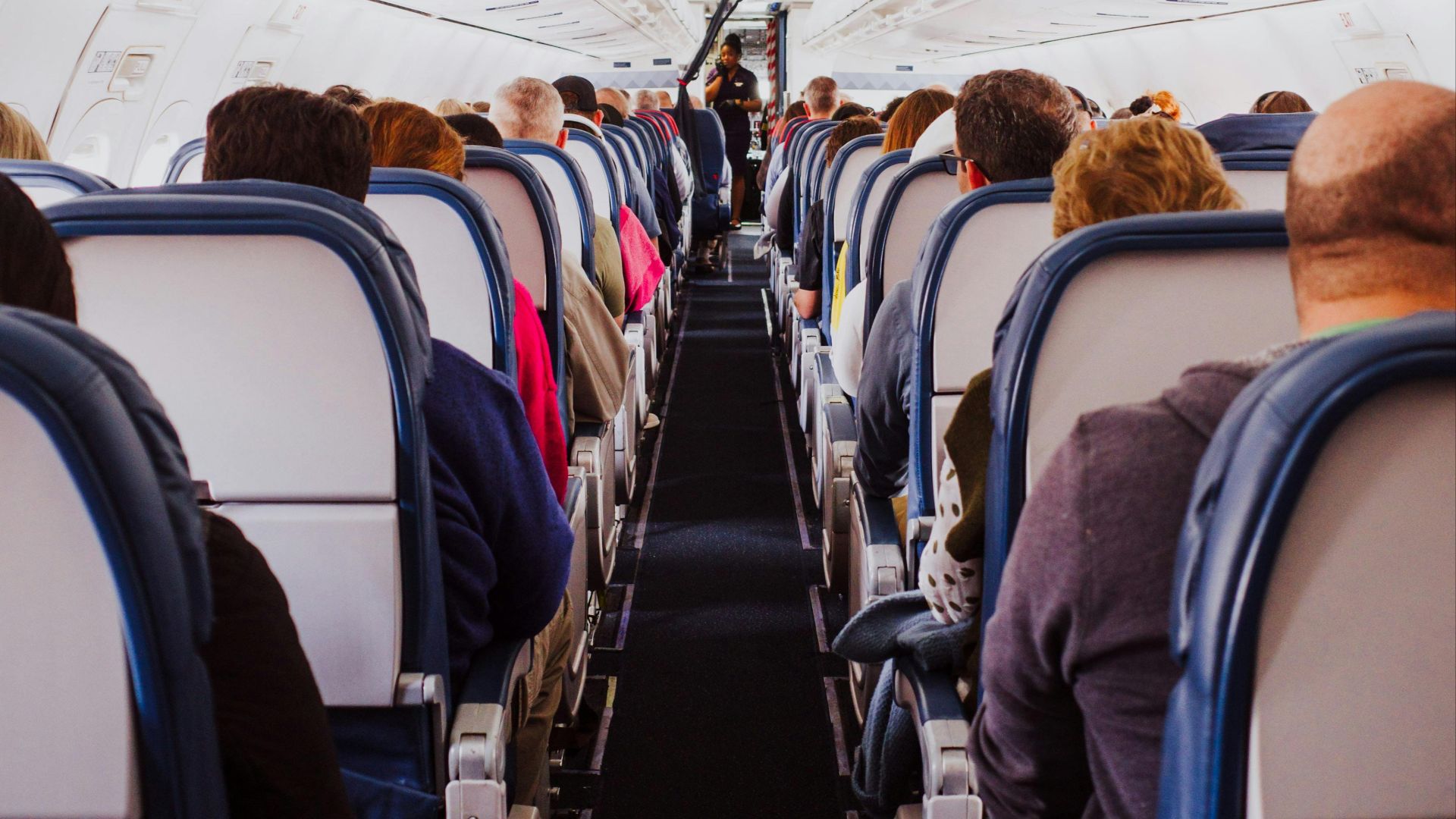View from the back of a crowded airplane cabin showing passengers seated in rows during flight.