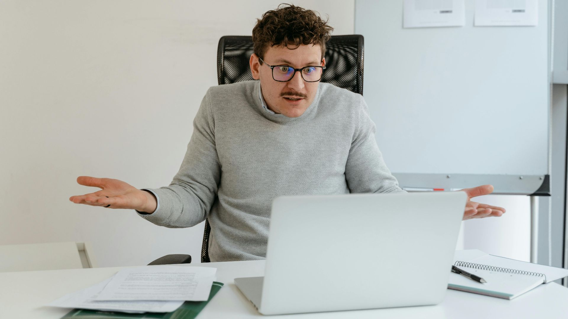 A man in a gray sweater and glasses gesturing at his laptop screen during a video call in an office setting.