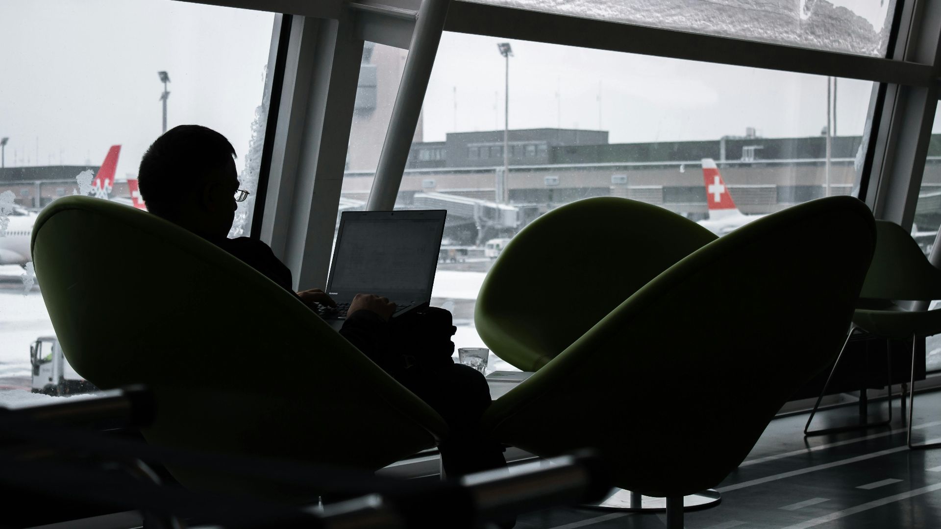 Silhouette of a man working on laptop at airport lounge with planes outside.