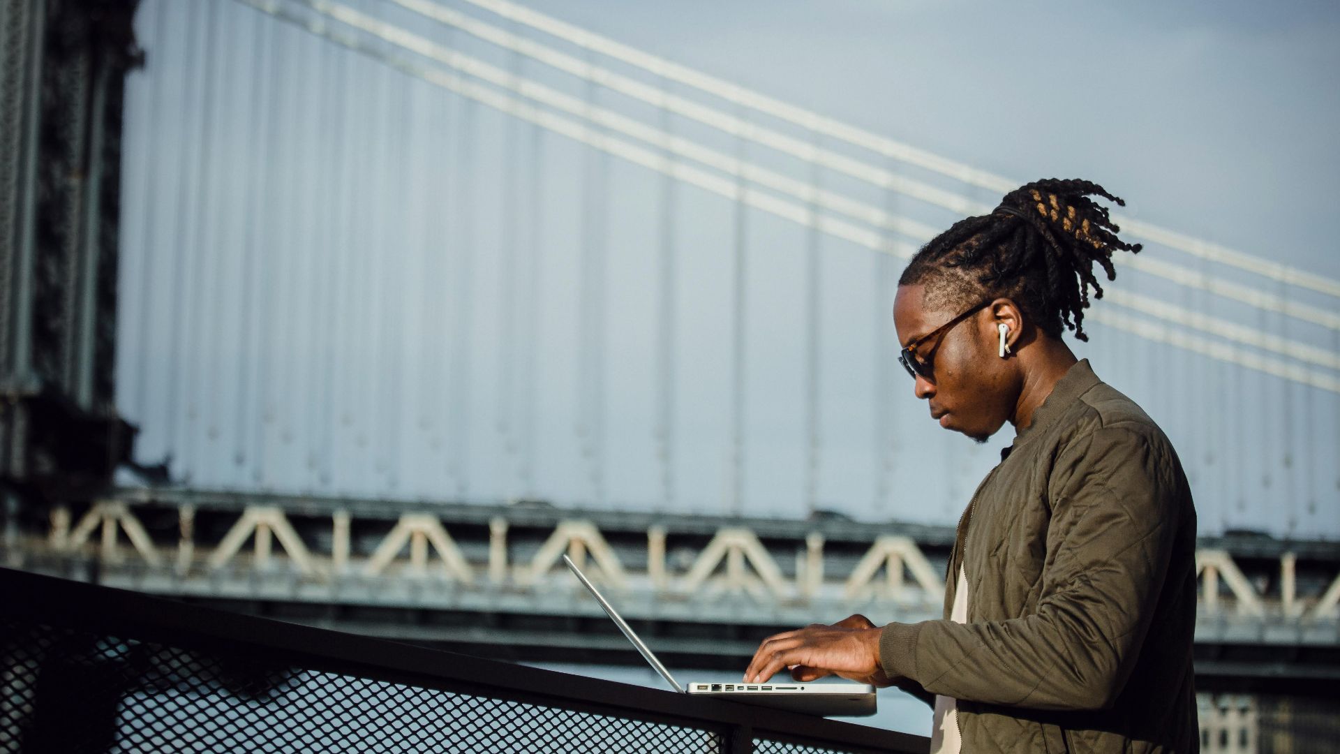Side view of cool young African American man with dreadlocks in sunglasses and earbuds working on laptop on terrace railing against Manhattan Bridge and blurred blue sky