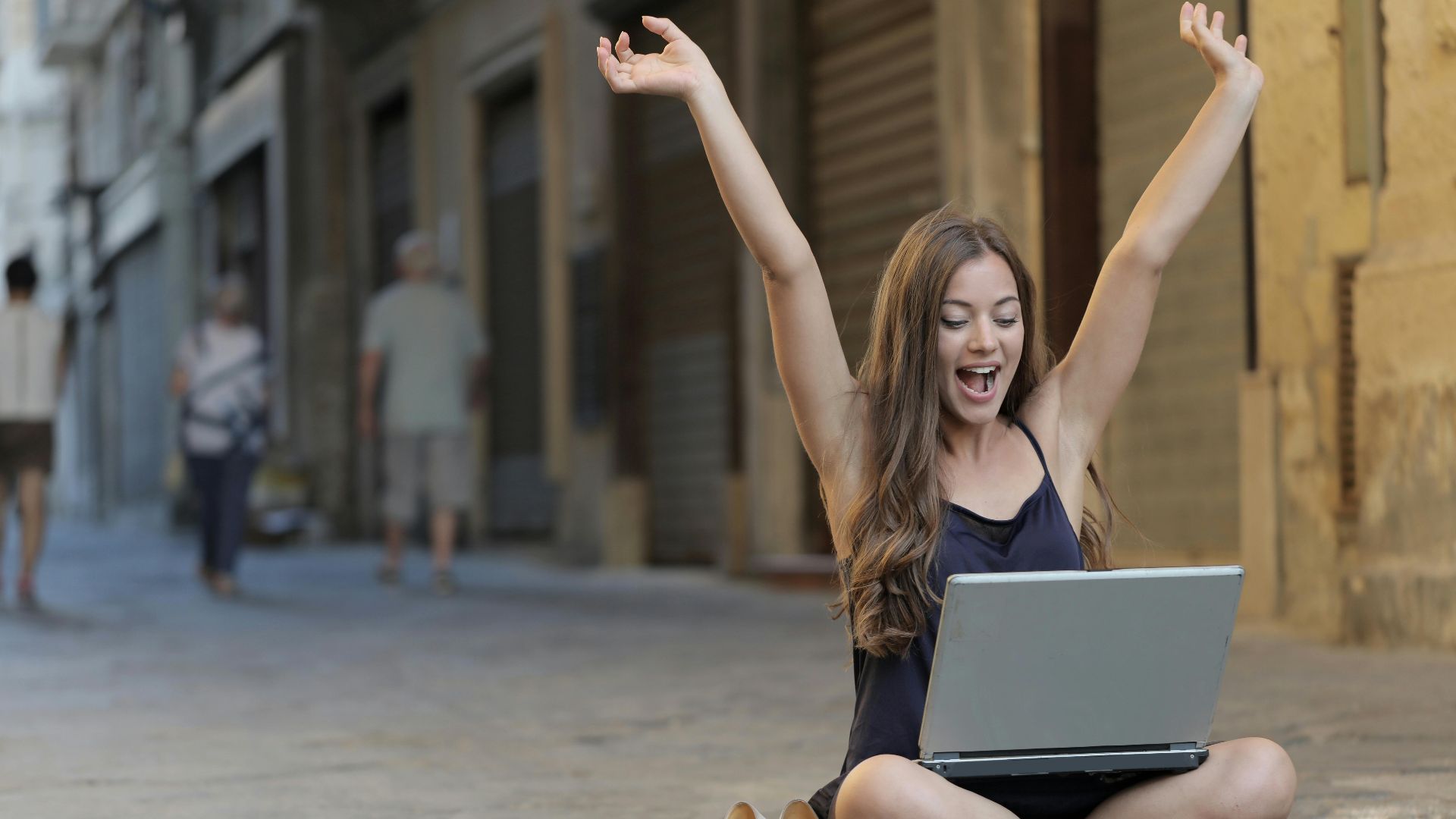 A cheerful woman sitting outdoors, celebrating success with arms raised, while using a laptop.