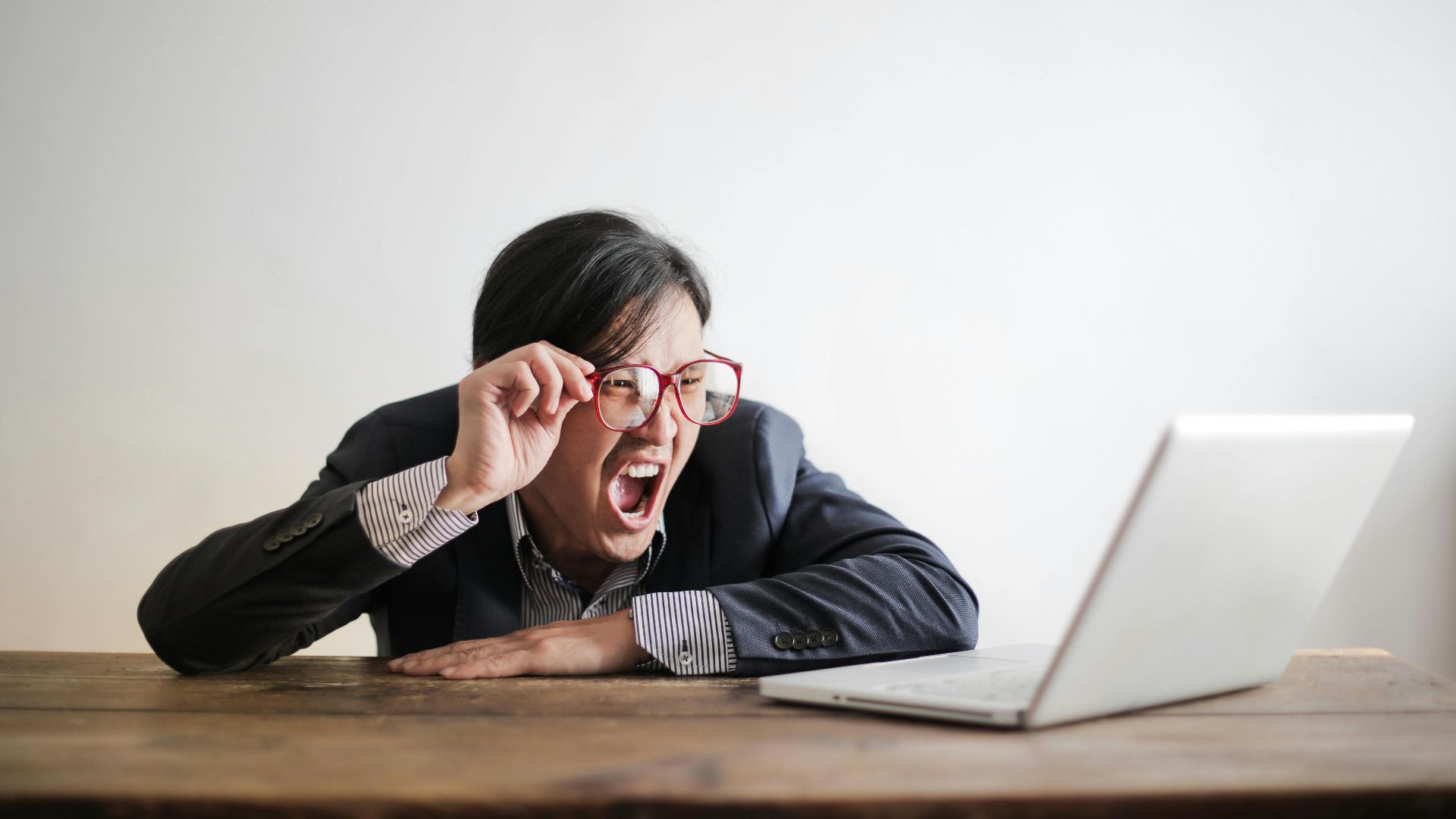 Modern Asian man in jacket and glasses looking at laptop and screaming with mouth wide opened on white background