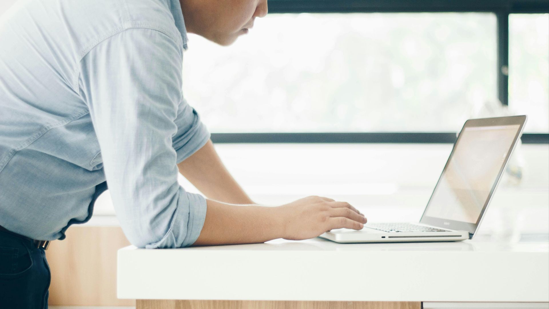 A man leaning on a desk working on a laptop in a bright indoor office setting.