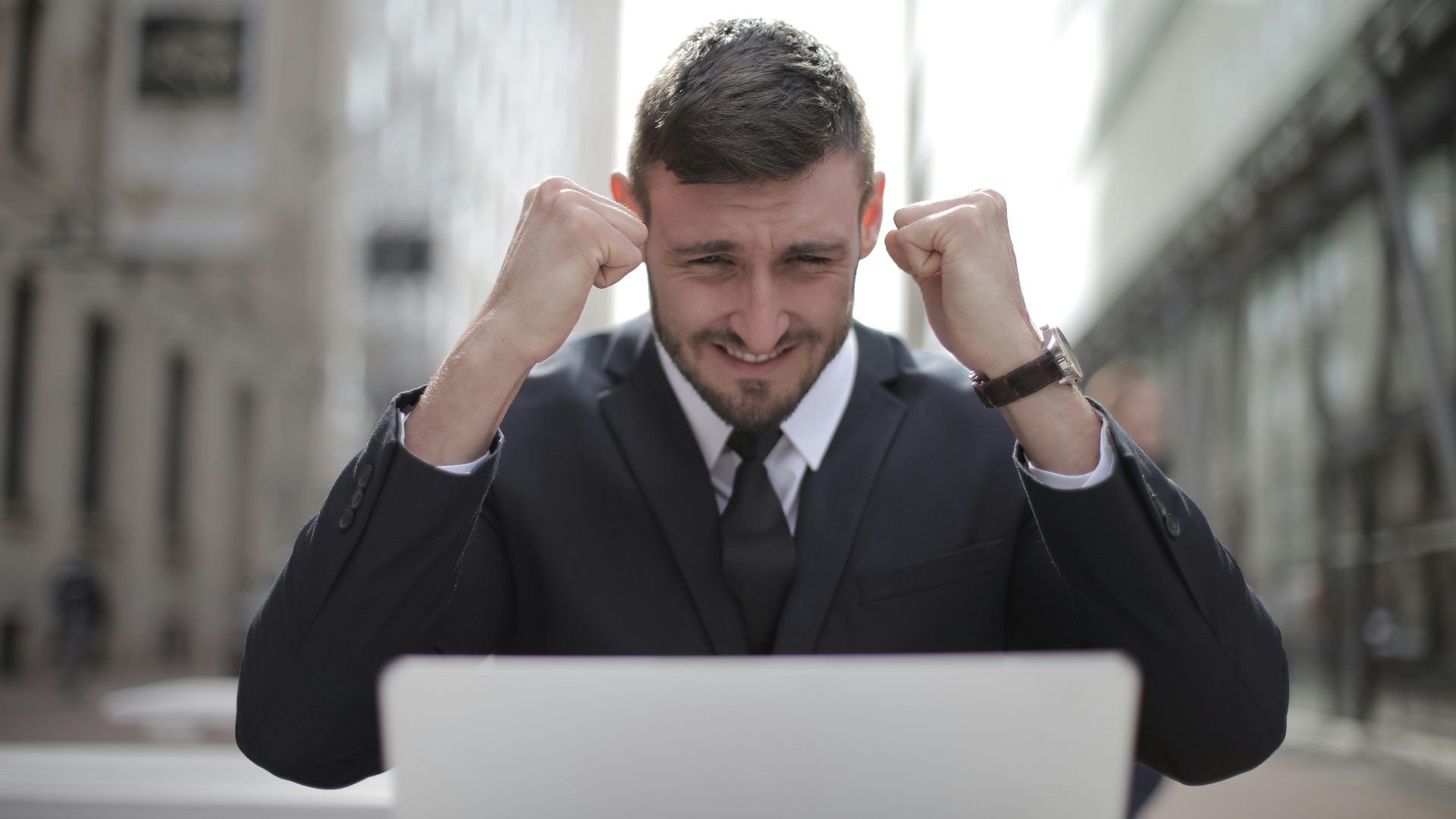 A joyful businessman in a suit triumphantly celebrates success on his laptop outdoors.