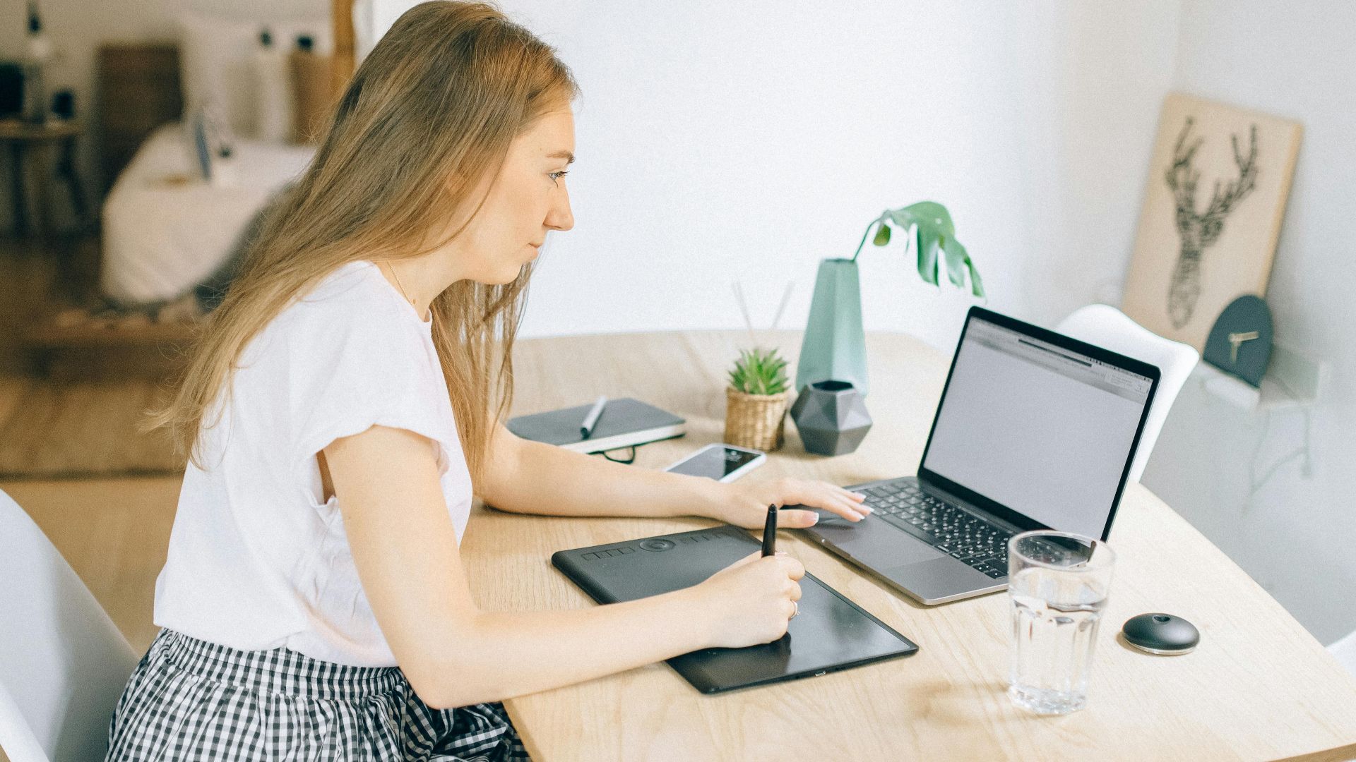 A young woman working remotely at a wooden table with her laptop and tablet in a cozy home setting.