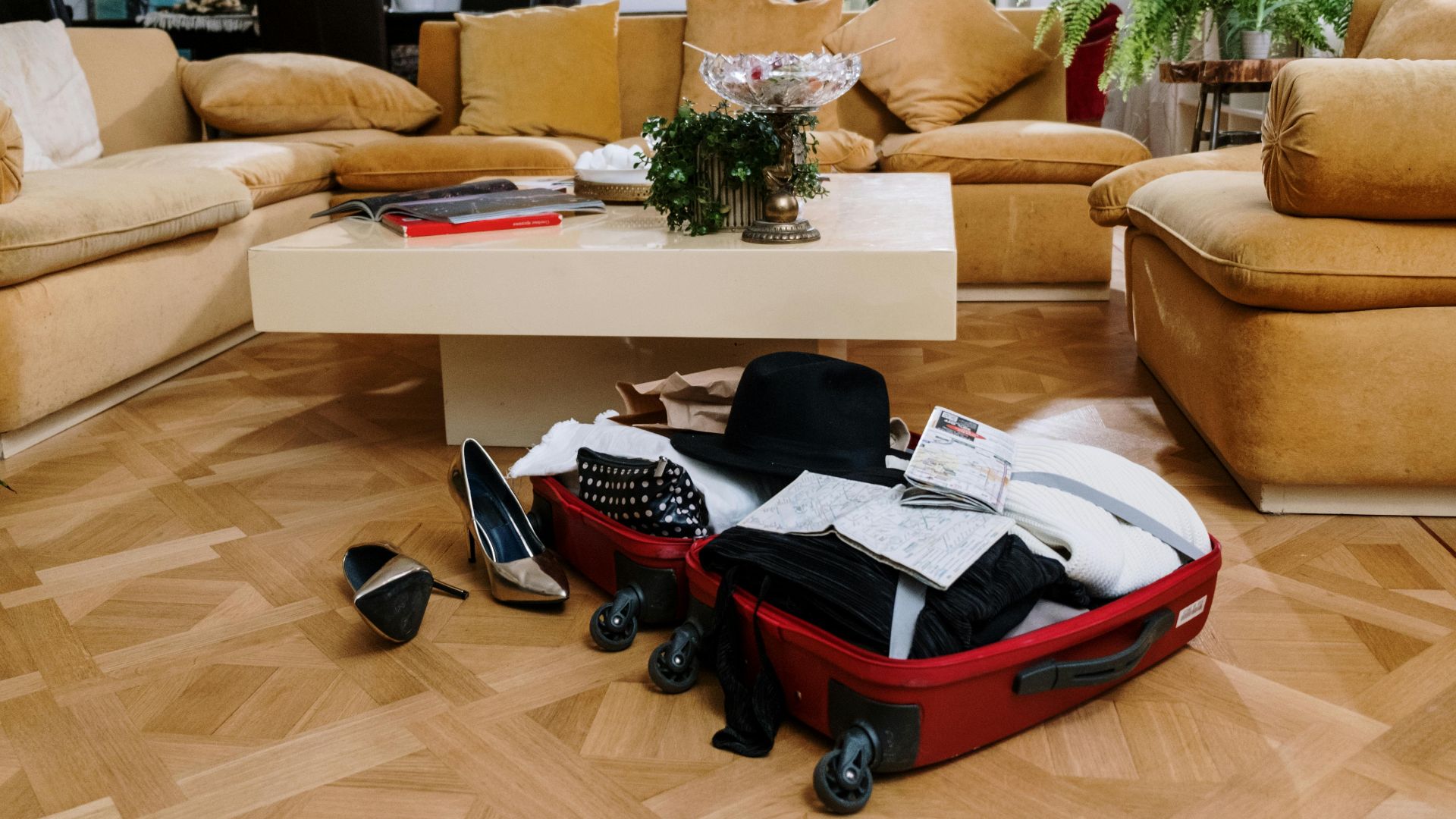 A stylish living room with beige couches and an open suitcase on a parquet floor, surrounded by plants.
