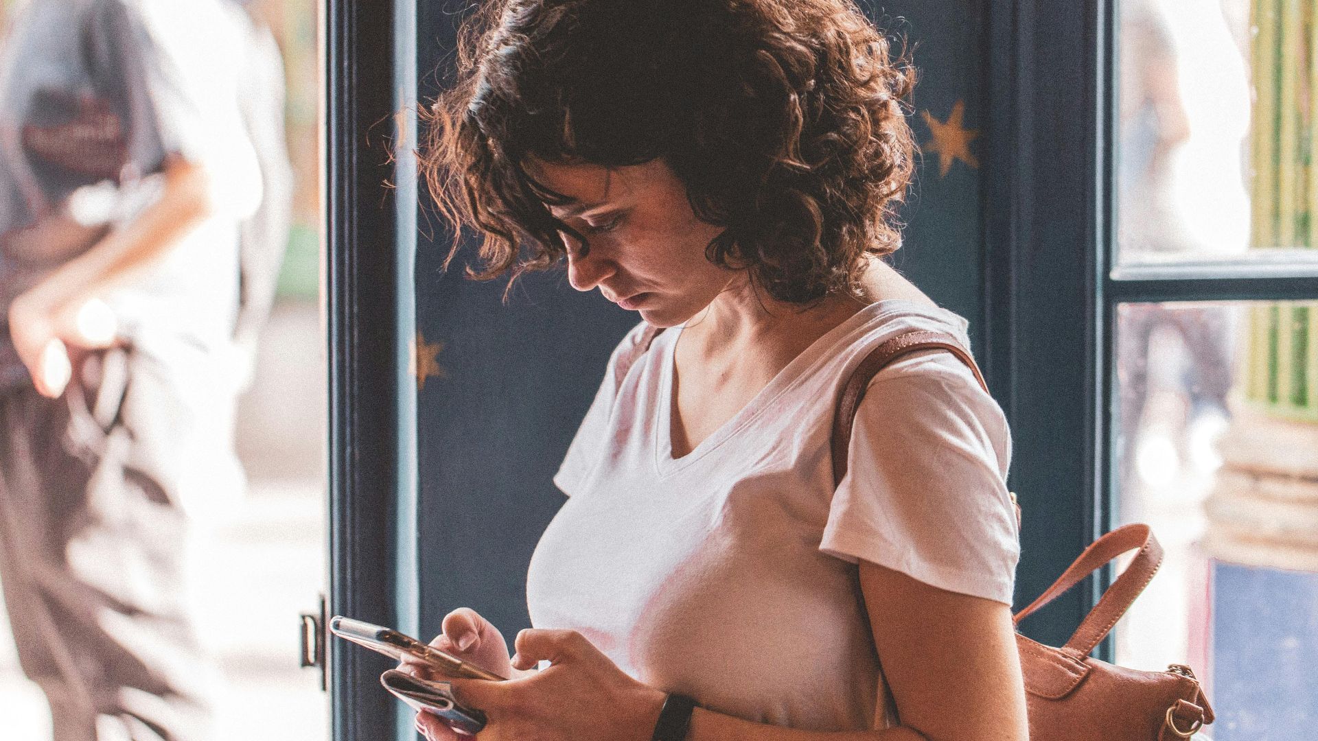 Woman with curly hair using smartphone by a sunlit window, wearing a white t-shirt.