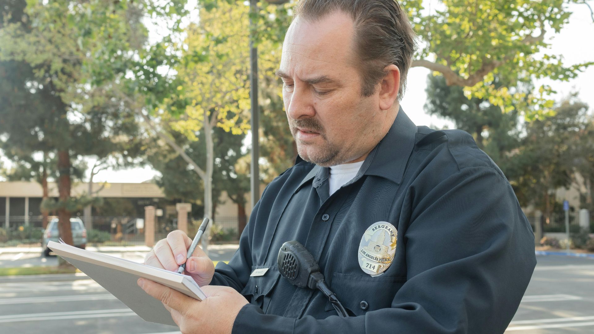 A police officer outdoors writing a report on a notepad. Trees in the background.