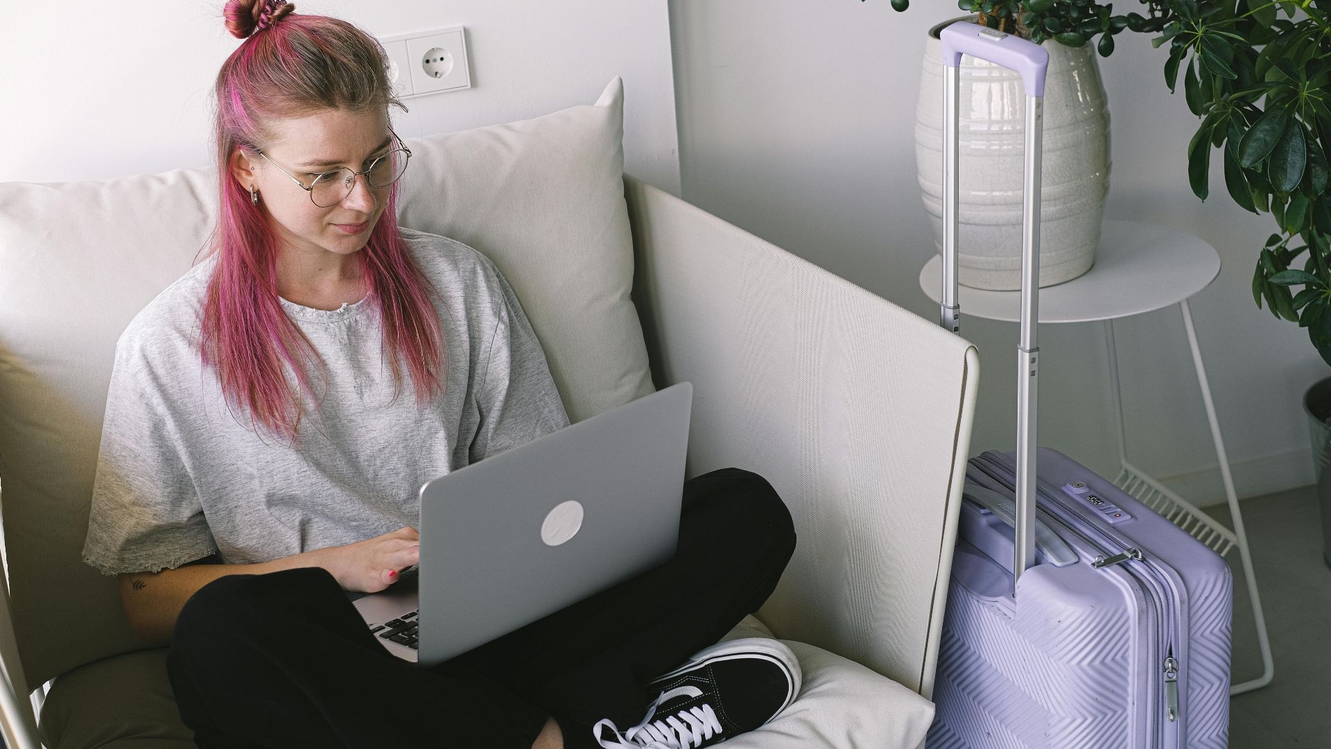 Casual young woman with pink hair works on laptop in comfortable indoor setting, travel suitcase nearby.