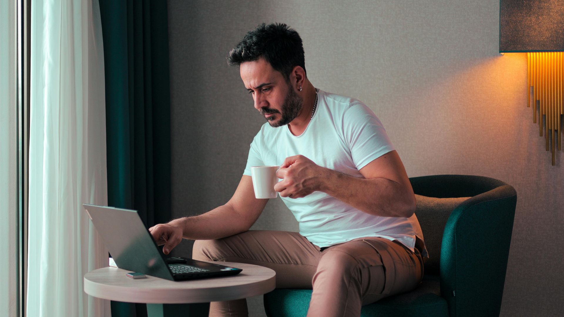 A man sits in a cozy hotel room in Cyprus, working on a laptop while enjoying a coffee.