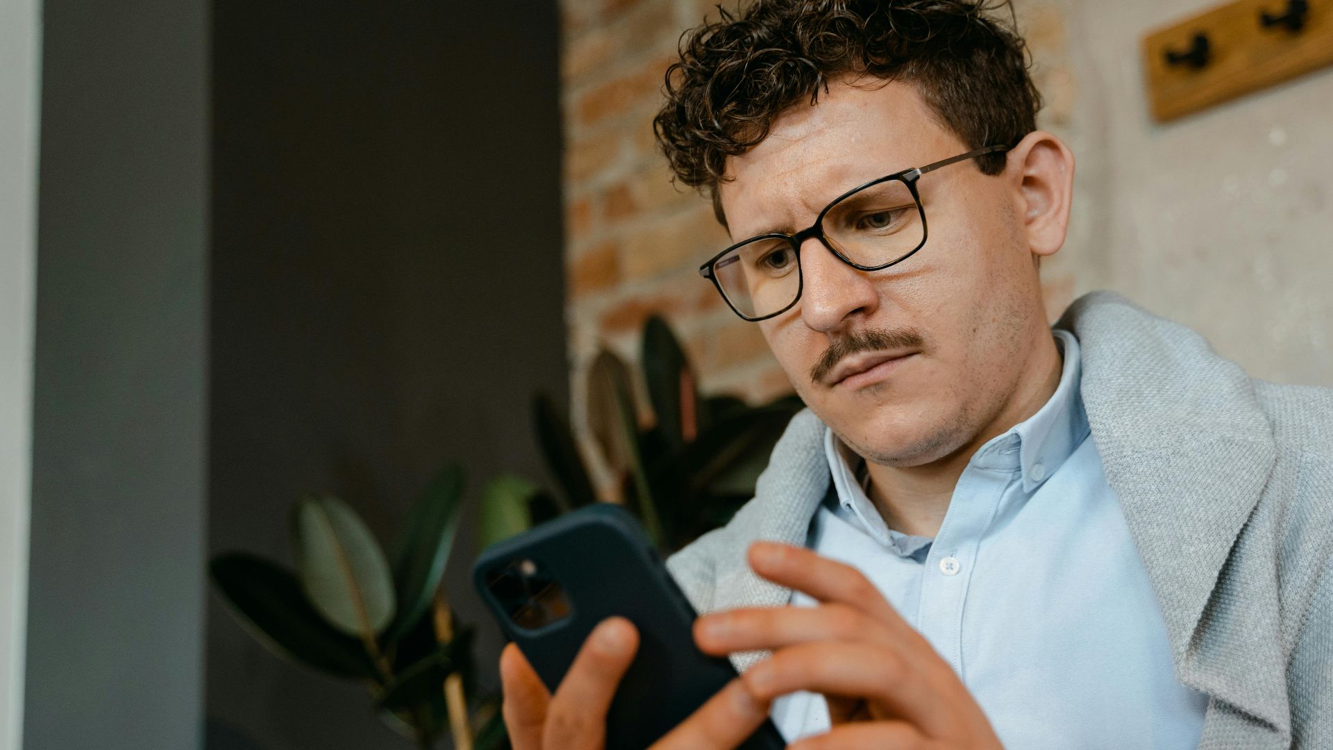 A focused man with glasses using a smartphone indoors next to a brick wall.