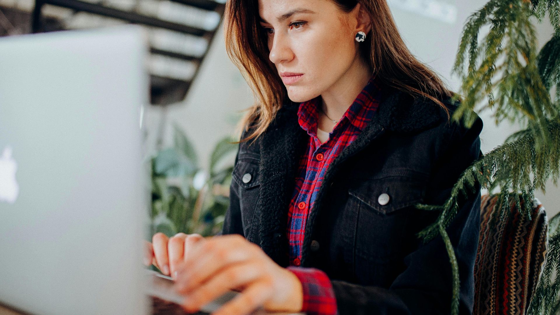 Young woman in plaid shirt intensely working on laptop in a modern environment with greenery.