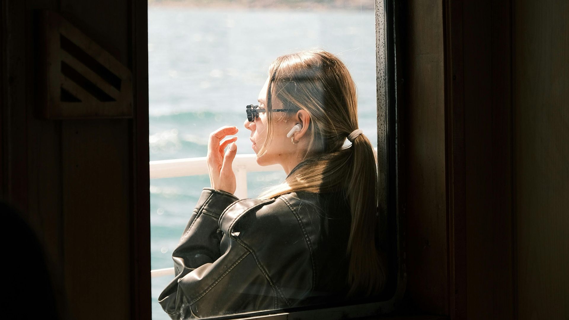 Woman wearing sunglasses, gazing through ferry window at scenic sea view.