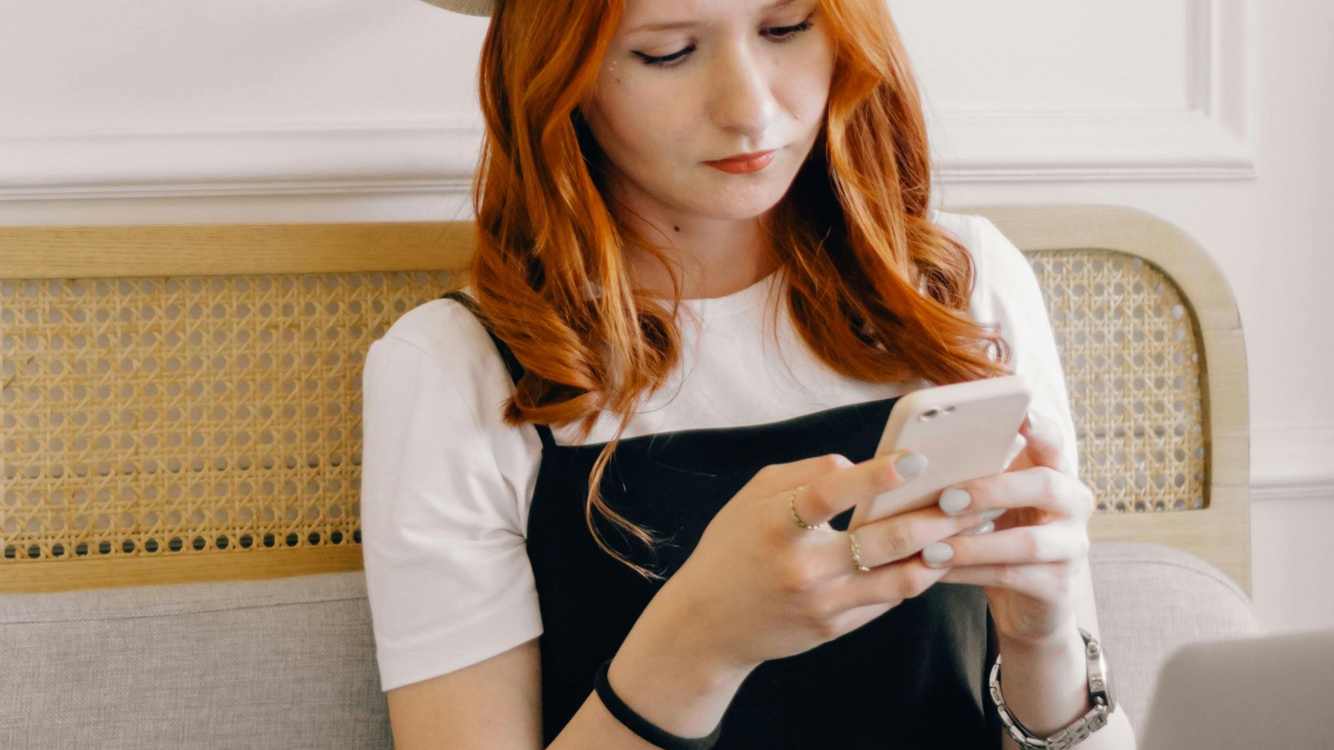 A young woman in a fedora hat texting on her smartphone while sitting with a laptop indoors.