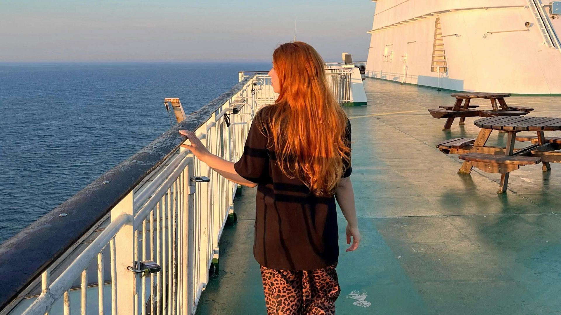 A woman with long hair walks on a cruise ship deck under a stunning sunset.