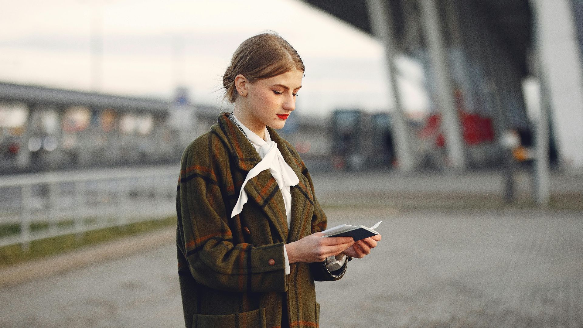 Attentive female passenger wearing trendy plaid coat and white blouse checking passport and ticket standing on pavement near modern building of airport outside