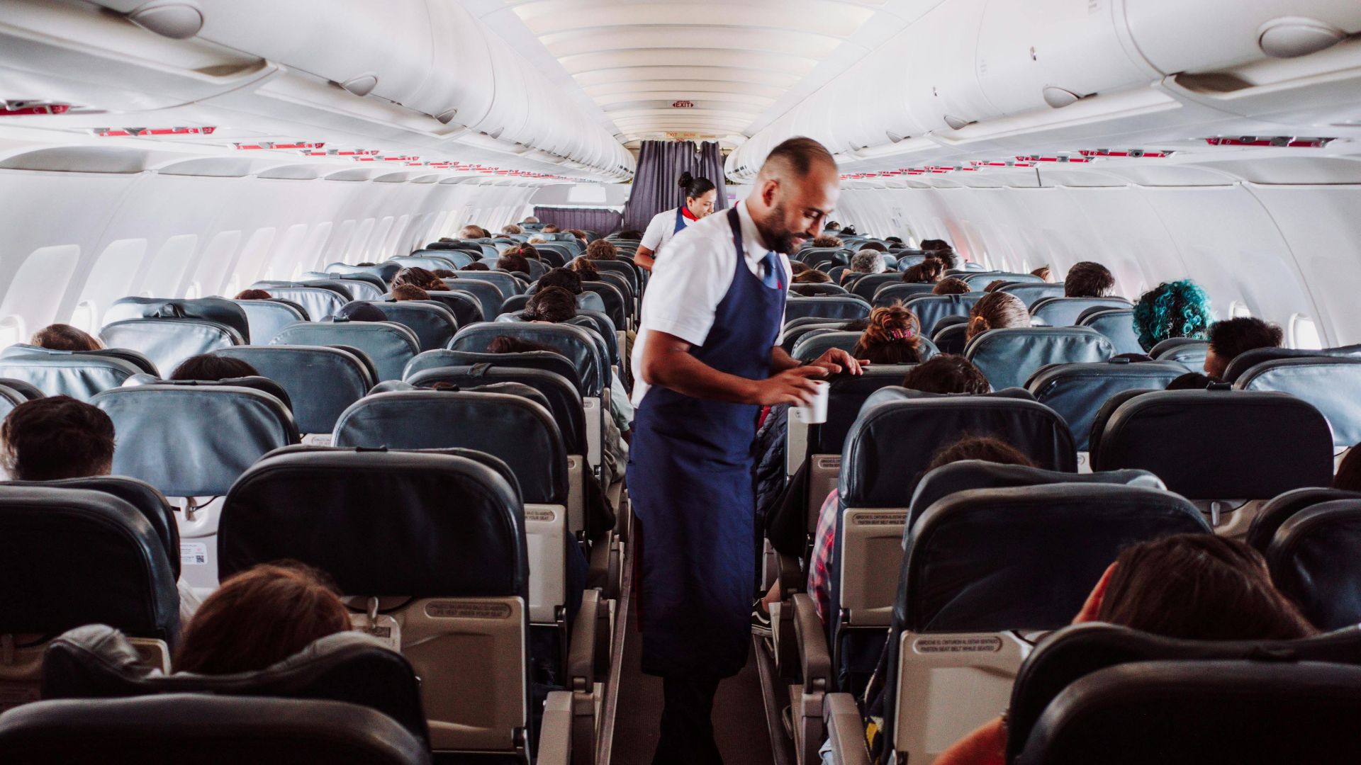 Flight attendant assists passengers in a crowded airplane aisle with rows of seats.