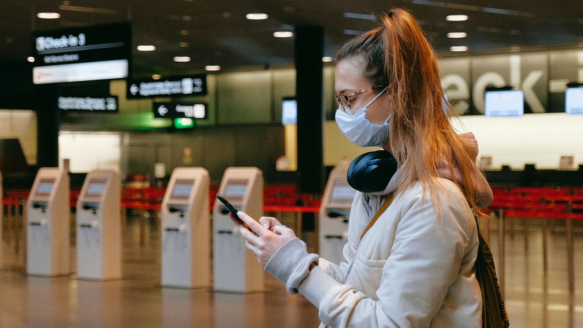 Woman wearing a mask, texting in airport terminal with signage in background.