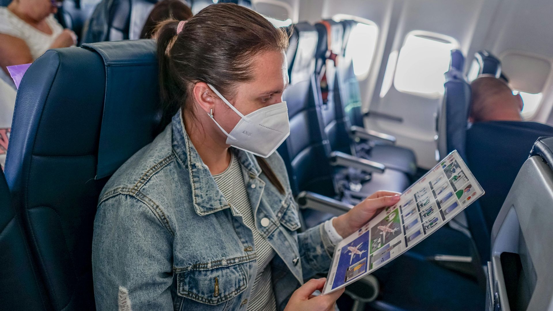 A woman wearing a mask on an airplane seat reading safety instructions.
