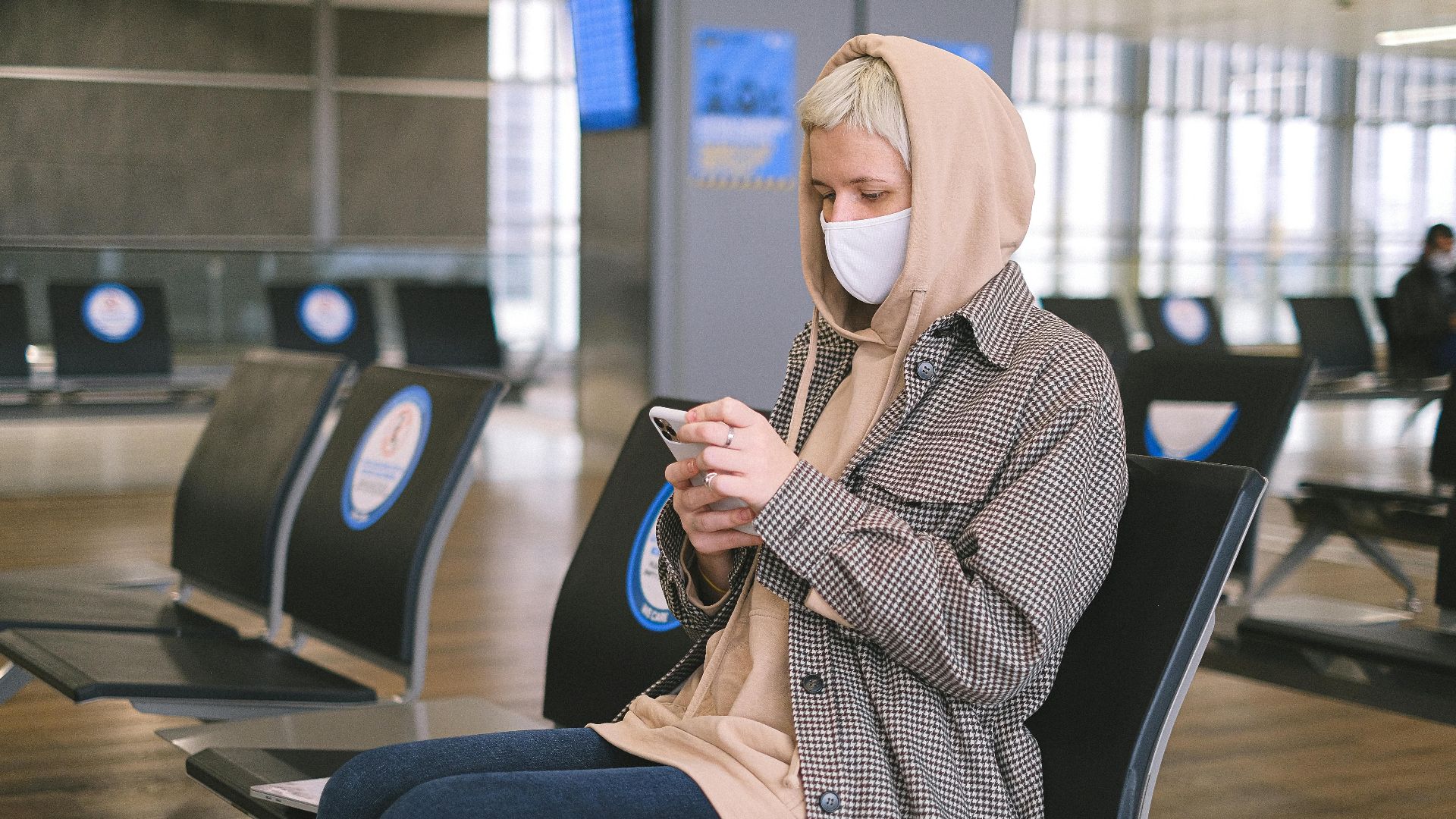 A woman wearing a face mask and a hood sits in an airport waiting area, using her smartphone.