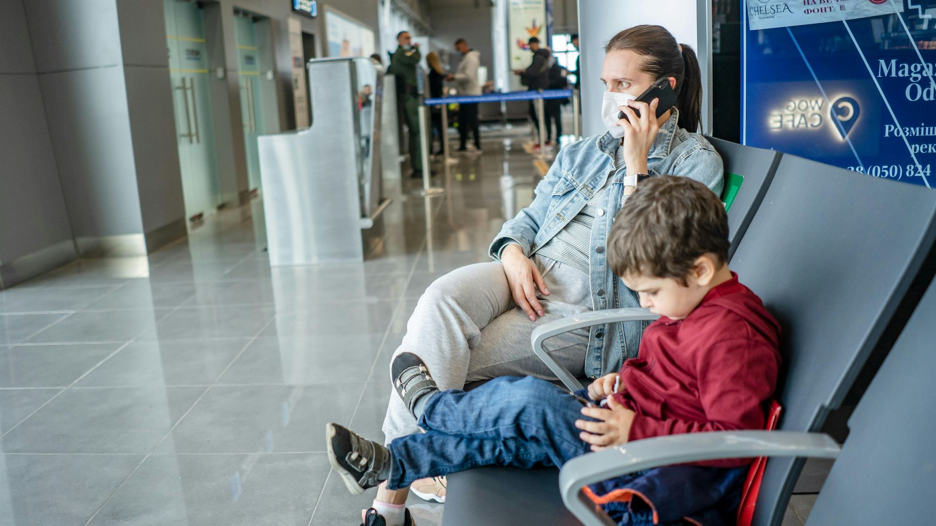 A mother and child sitting at an airport terminal. The mother is on the phone while the child is using a device.
