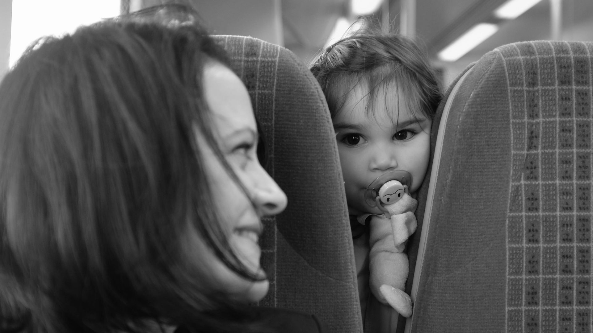 Mother smiling at her child playing peekaboo on a train, captured in black and white.