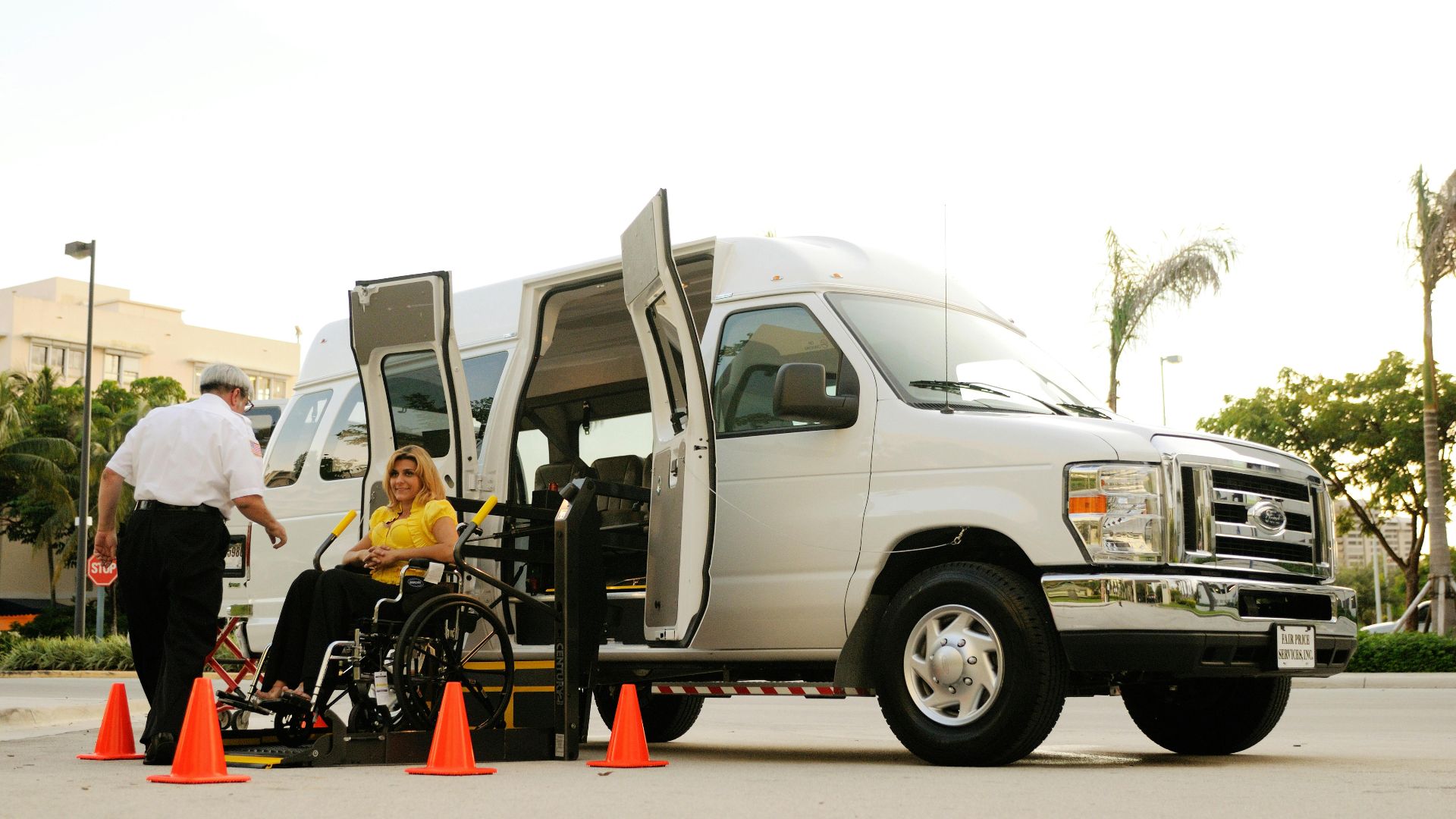 A person in a wheelchair uses a lift to board a van, assisted by a driver. Outdoor setting with cones.