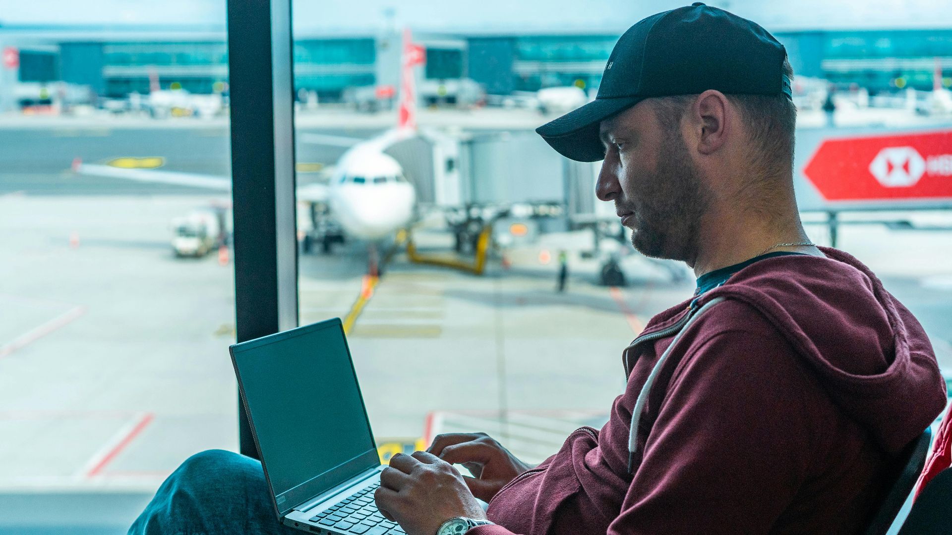 A man working on his laptop in an airport terminal, with a plane visible outside.
