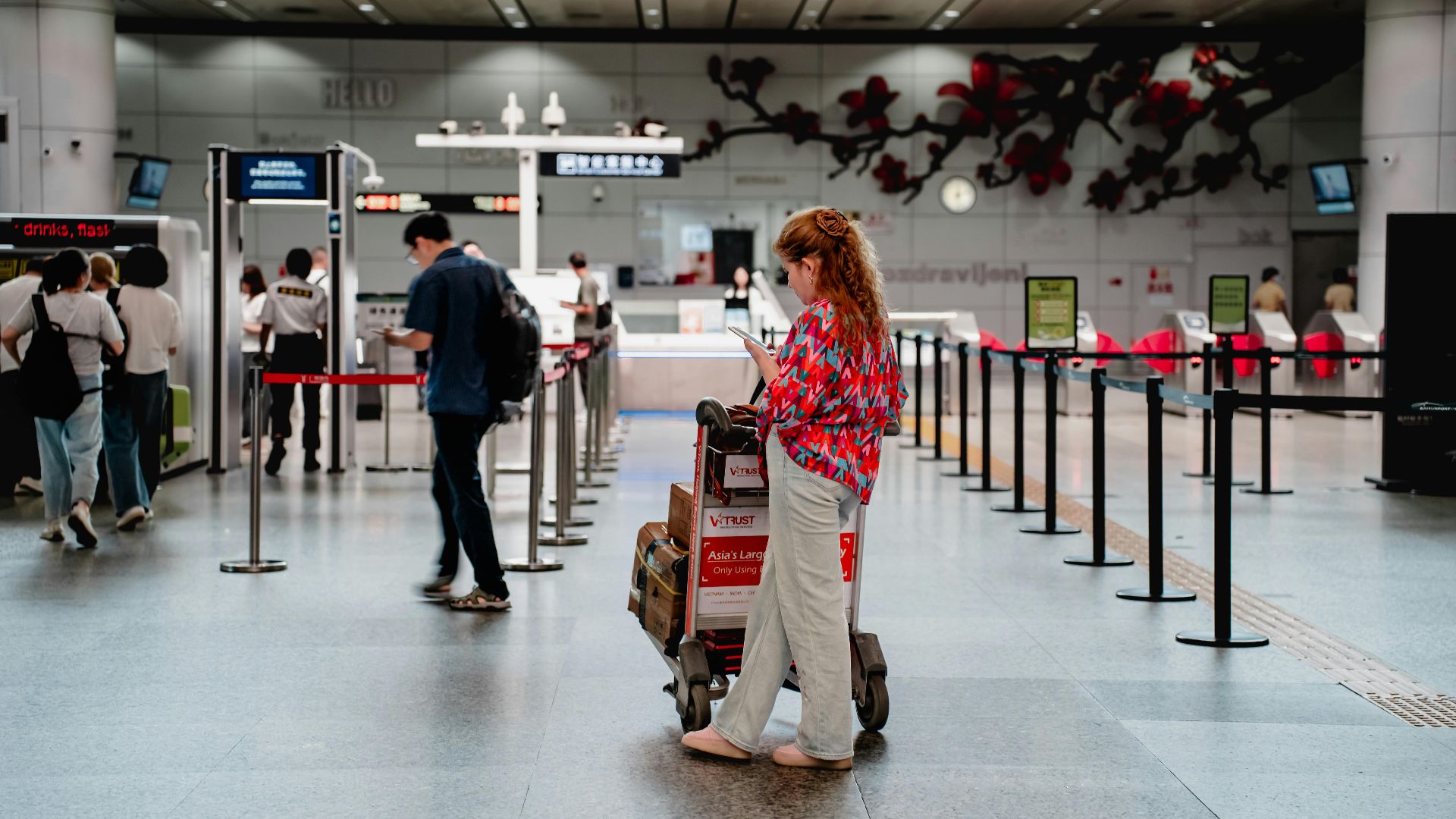 A traveler checks her phone while waiting with a luggage cart at a busy airport terminal in Guangzhou.