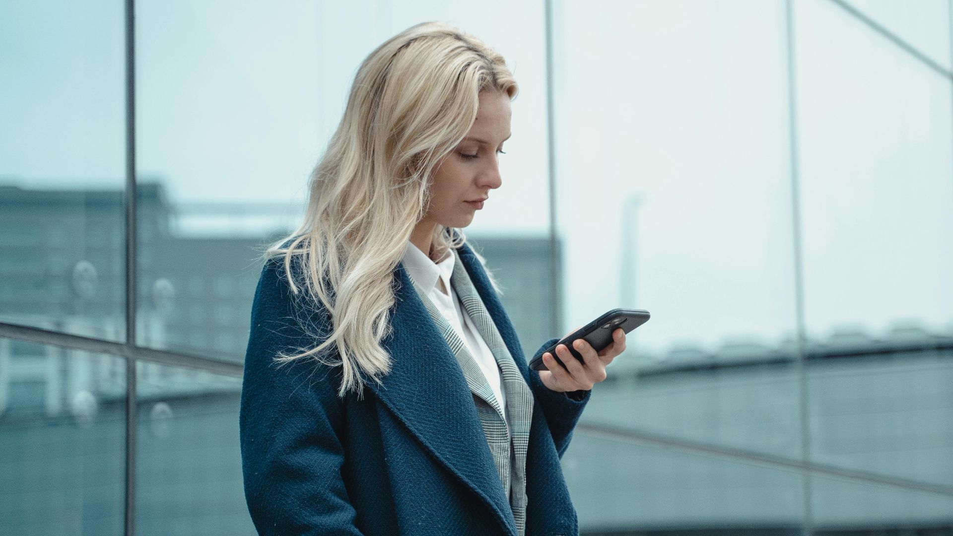 Professional woman in blue coat using smartphone outside office building.