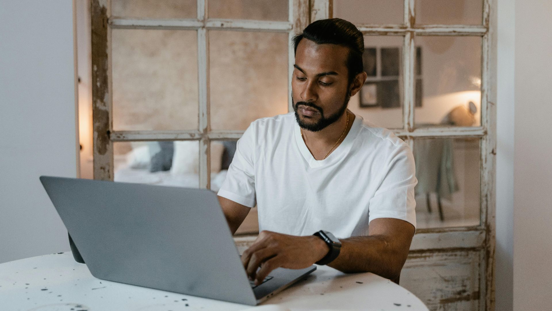Adult man with beard focused on laptop work at home desk during the day.