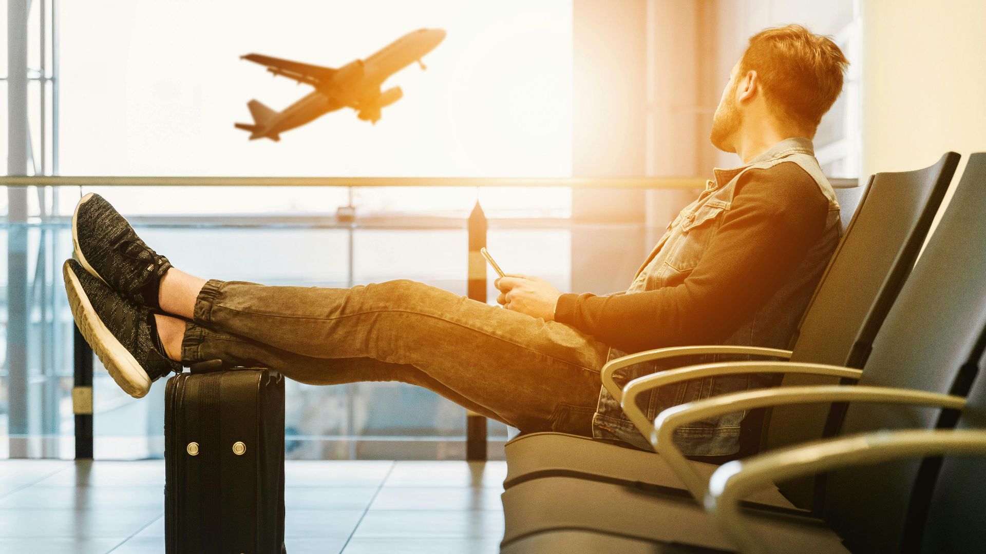 Side view full body of bearded man sitting on chair in waiting hall of airport and looking at flying aircraft behind window