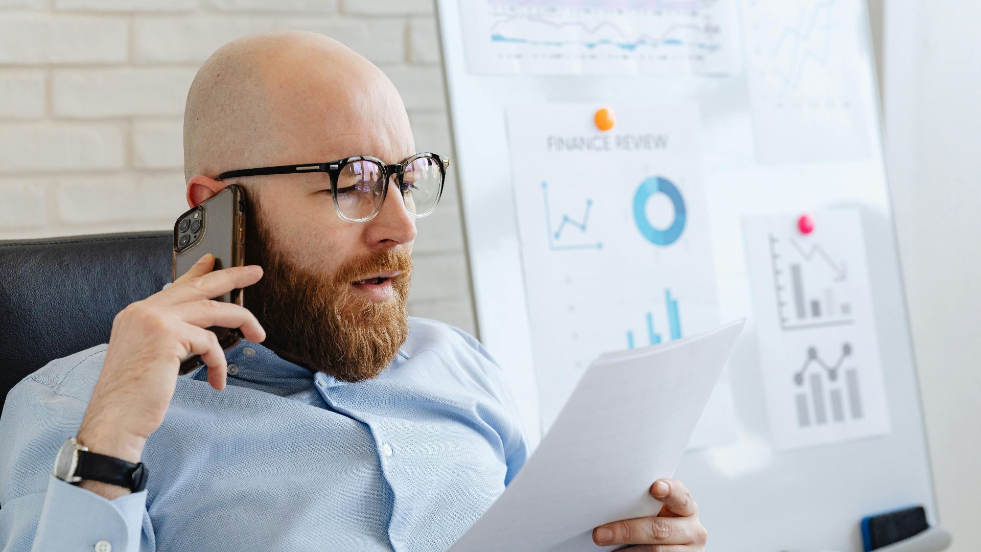 Bald man with beard holding smartphone and papers, reviewing financial charts in office.