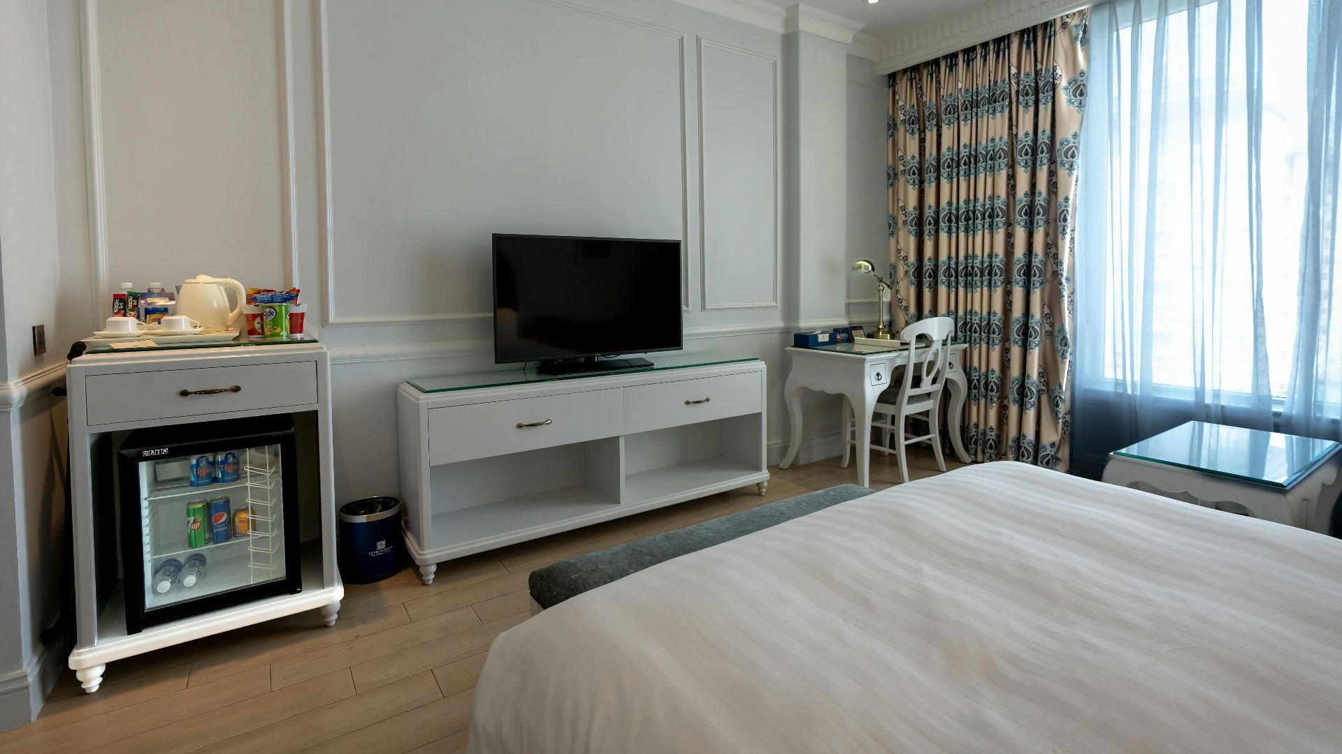 Bright and stylish hotel bedroom with TV, minibar, and cozy furnishings.