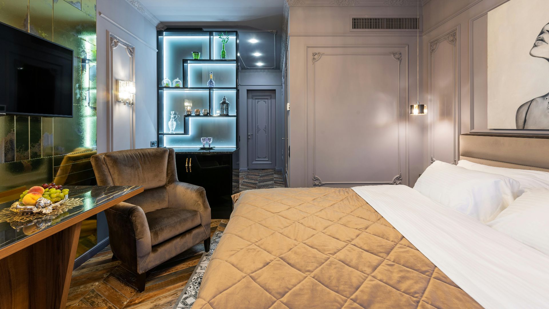 Table and armchair against bed with cover under glowing chandelier in house bedroom with entrance door