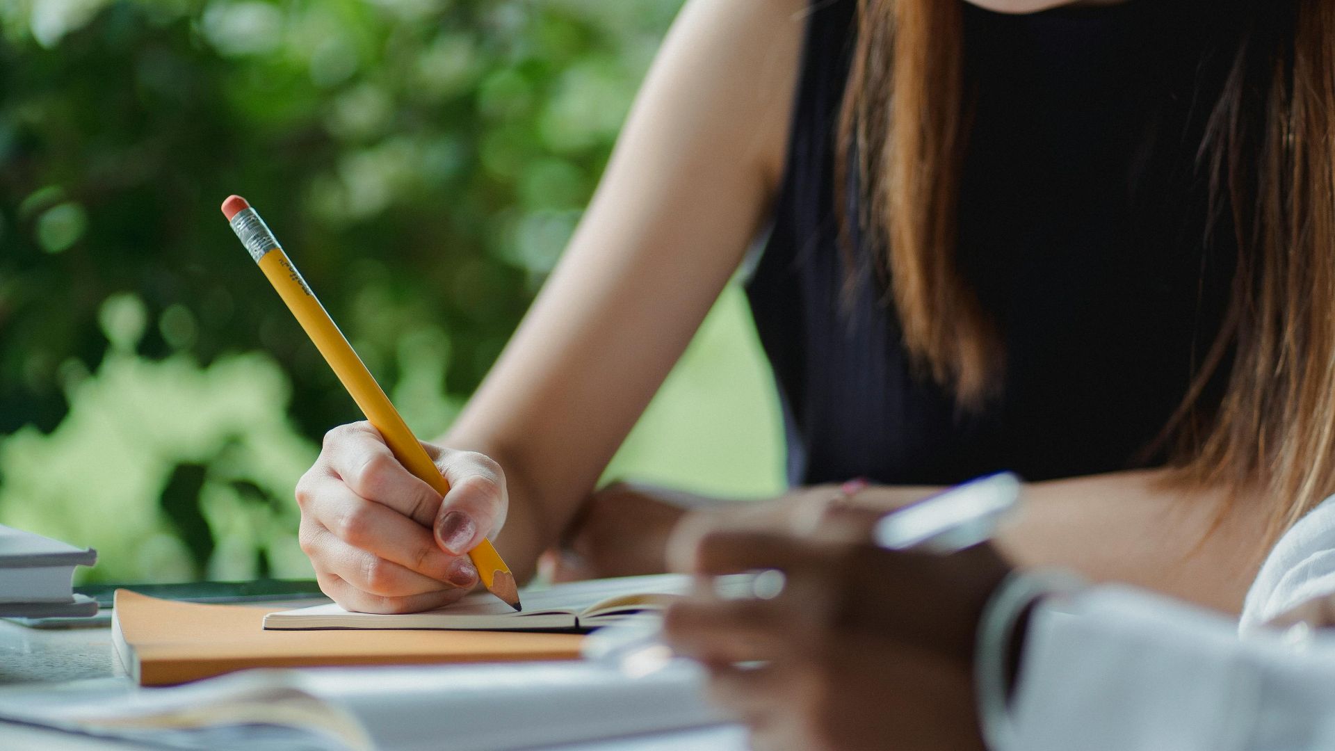 Concentrated Asian female student taking notes while doing research with African American female sittin at table