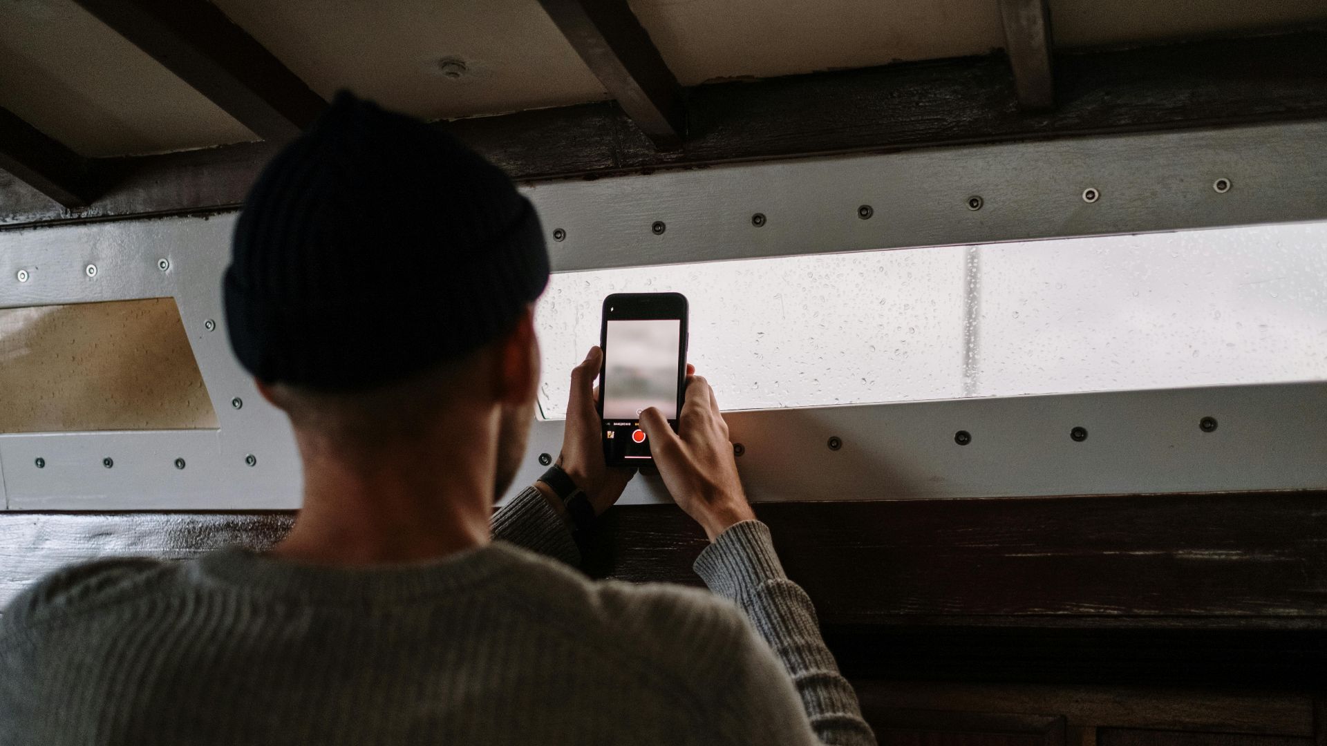 A man in a gray sweater takes a picture with his smartphone inside a wooden room.