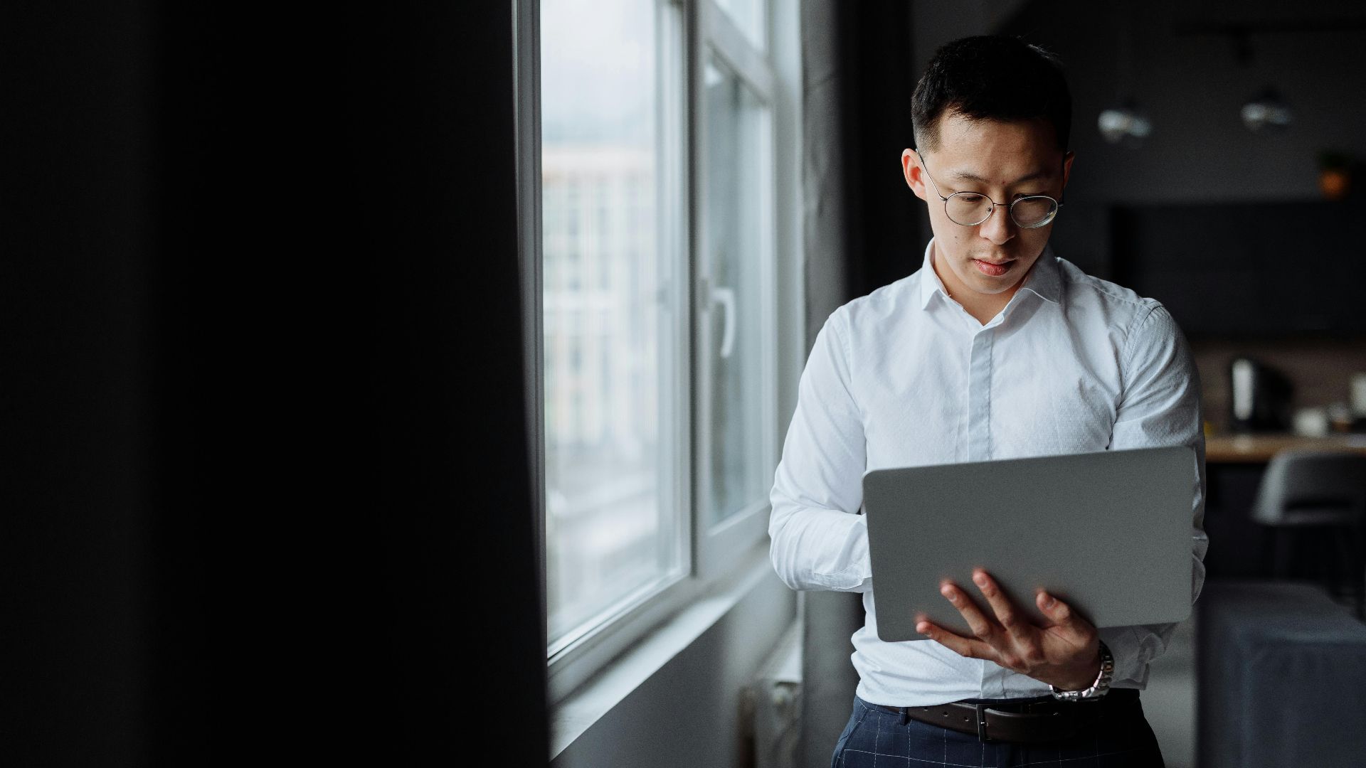 An Asian man in a white shirt using a laptop while standing by a window indoors.