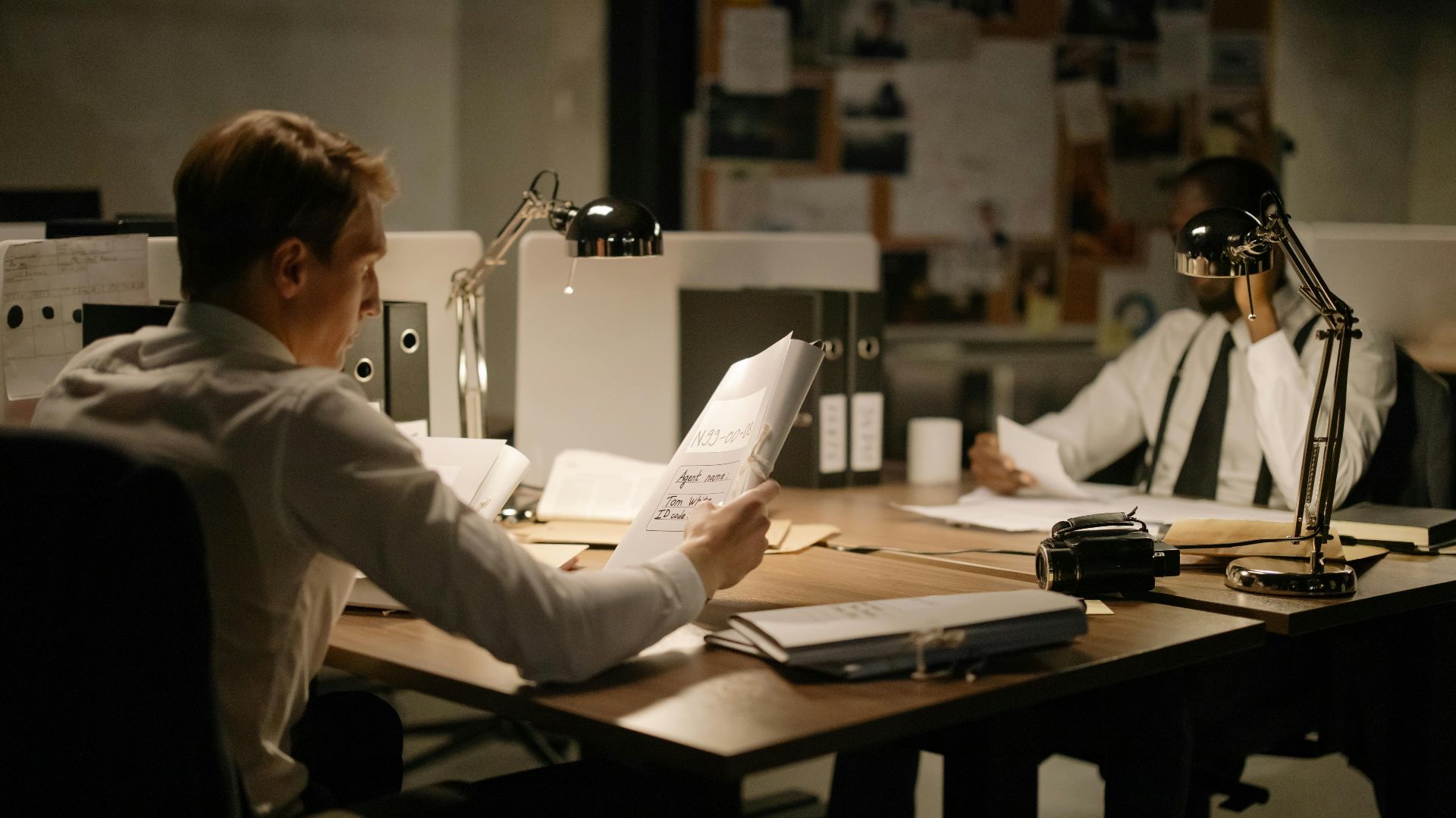Two detectives analyzing documents in an office setting under dim lighting, focusing on investigation.