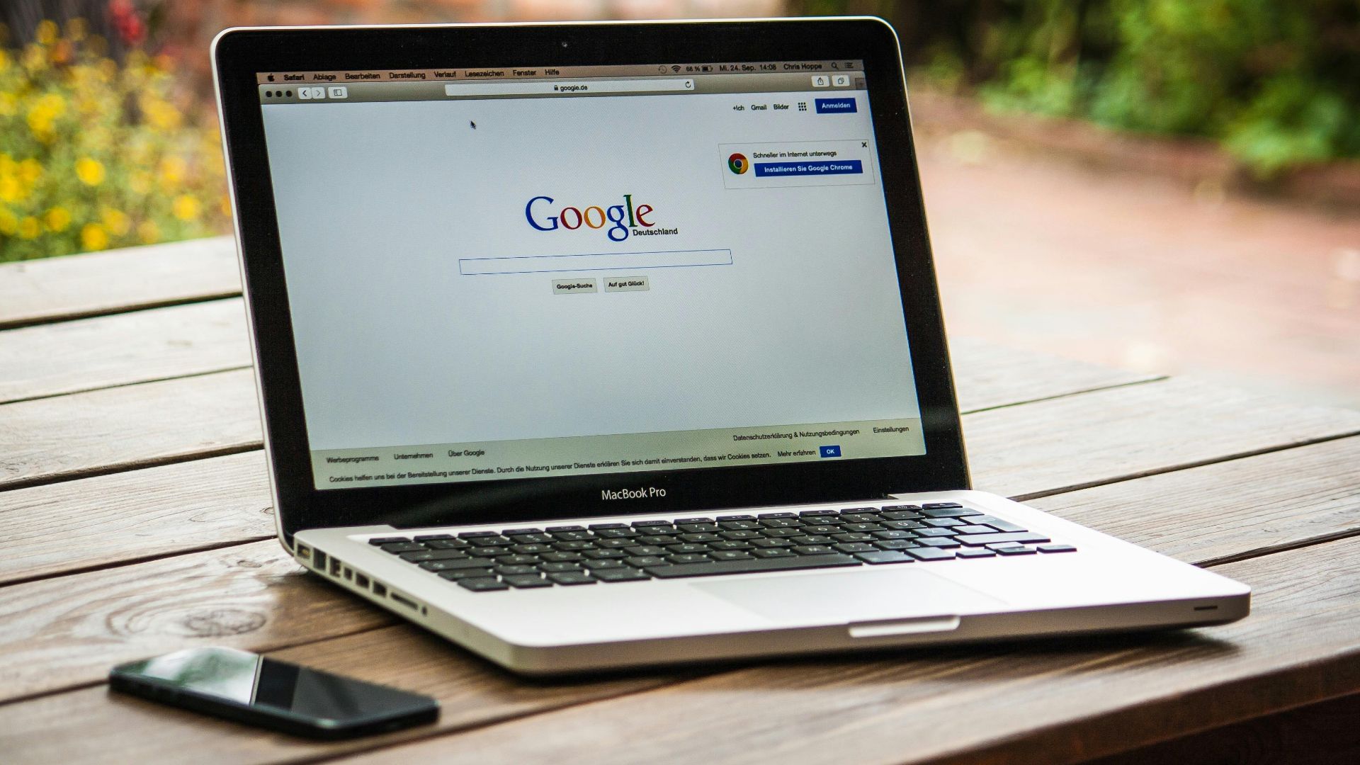 A MacBook Pro displaying Google Search on a wooden table outdoors, next to a smartphone.