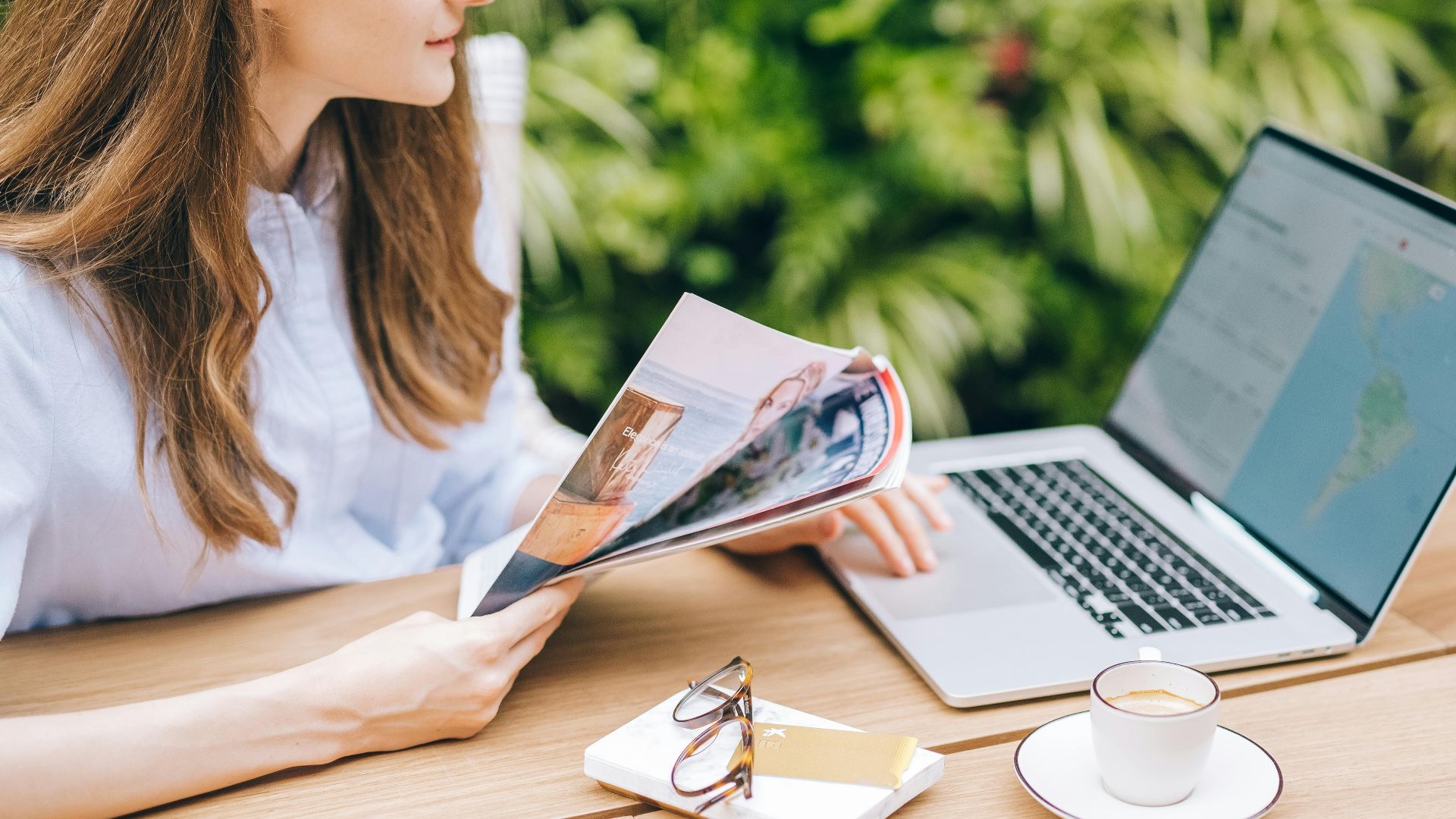 A woman sits outdoors, enjoying a magazine with coffee and laptop nearby.