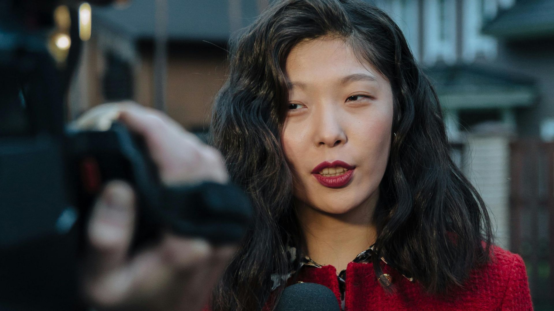 Asian woman reporting news on camera outdoors, wearing red coat.