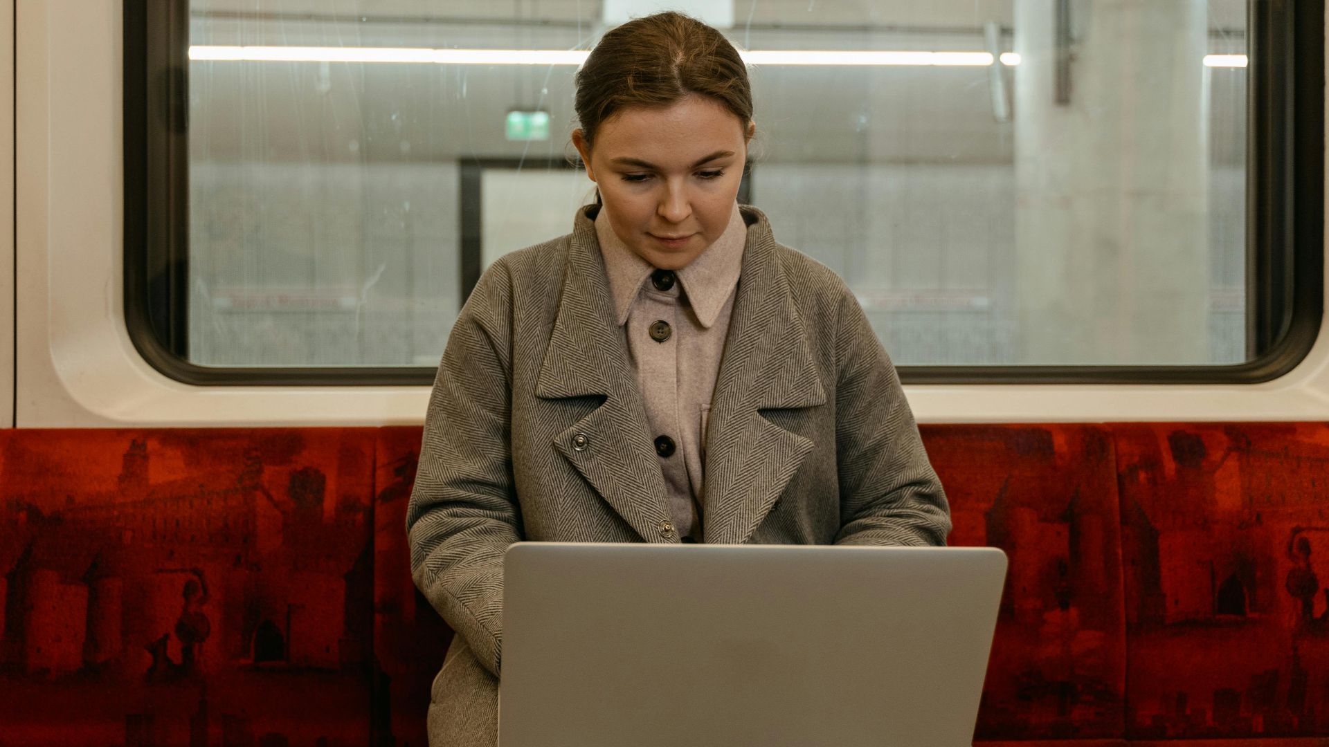 Caucasian woman using laptop in a gray coat sitting inside a subway train.
