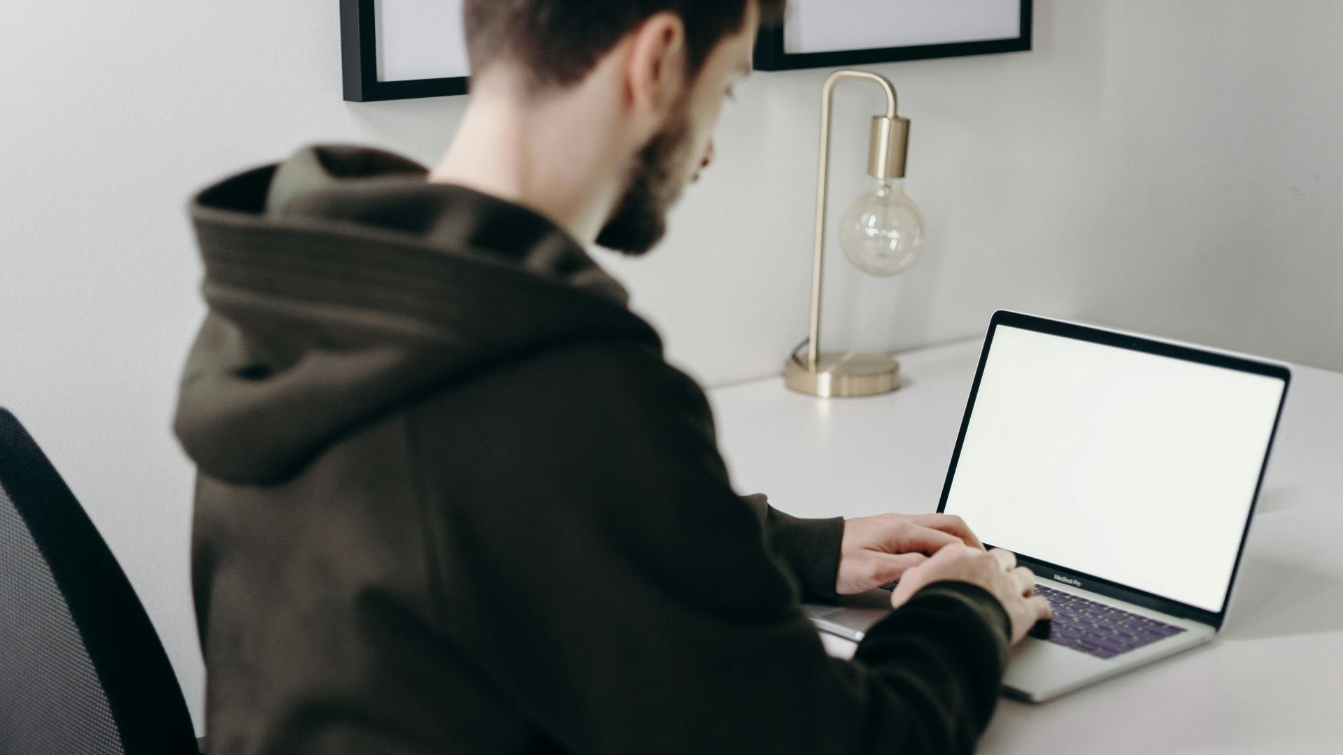 A young man in a hoodie working on a laptop at a minimalist home office desk.