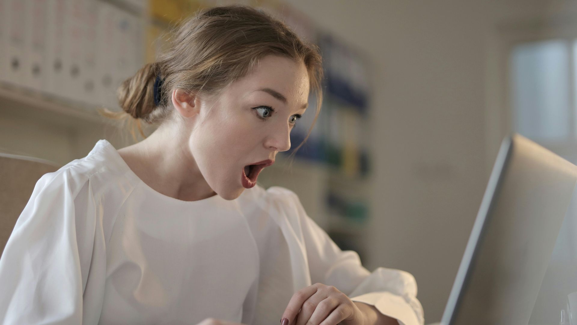 Surprised woman sitting at desk with laptop indoors, expressing amazement.