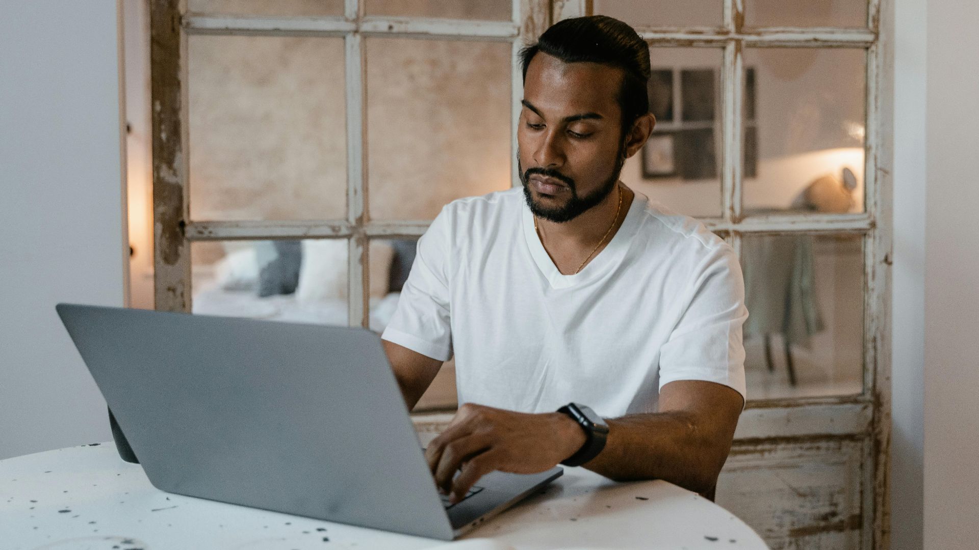 Adult man with beard focused on laptop work at home desk during the day.
