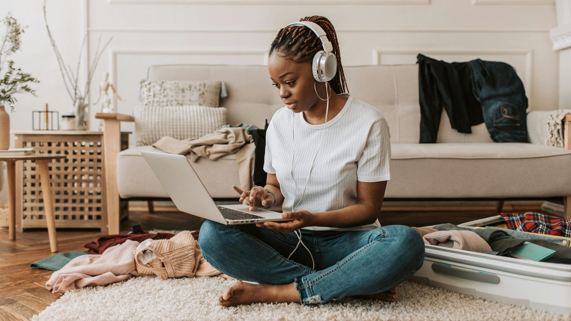 A young woman sits on the floor using a laptop and headphones, amidst clothes and a suitcase.