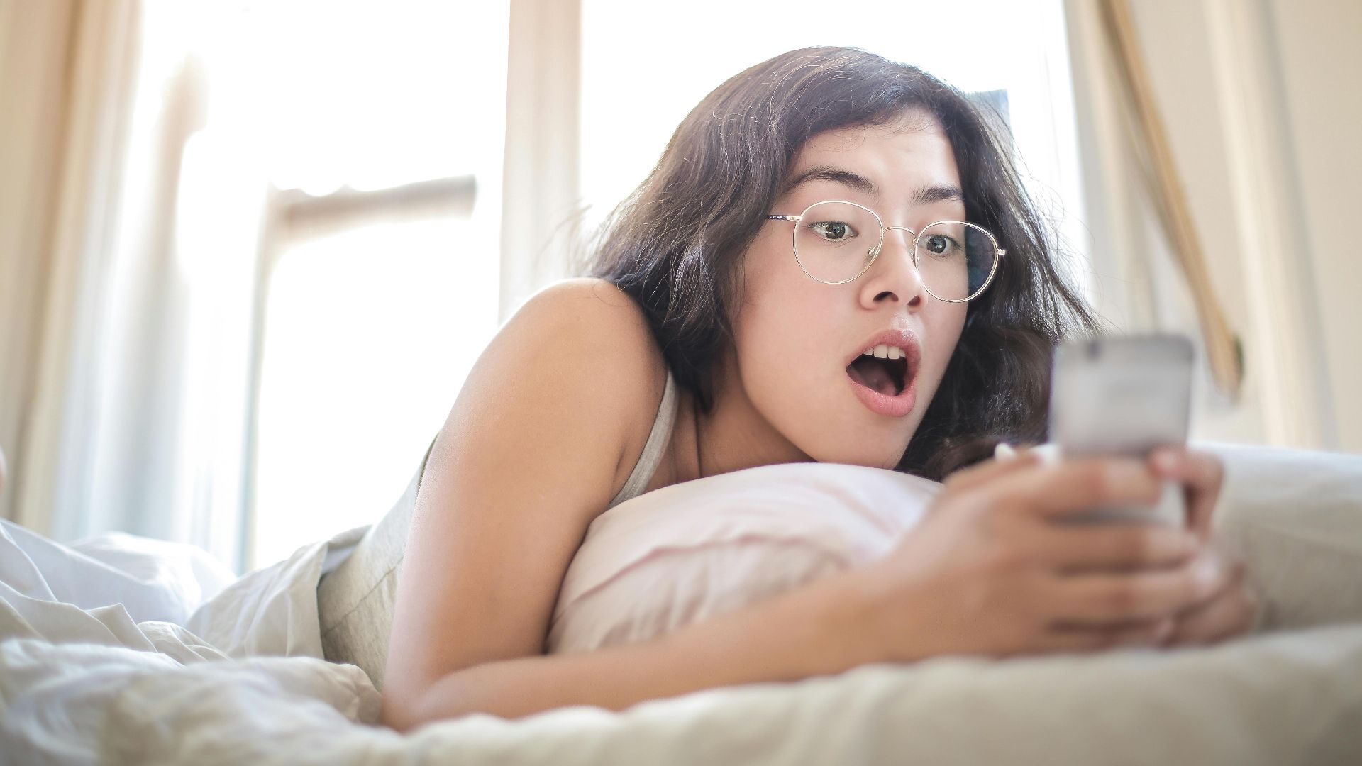Young woman lying on bed, surprised by something on her smartphone, in a bright bedroom setting.